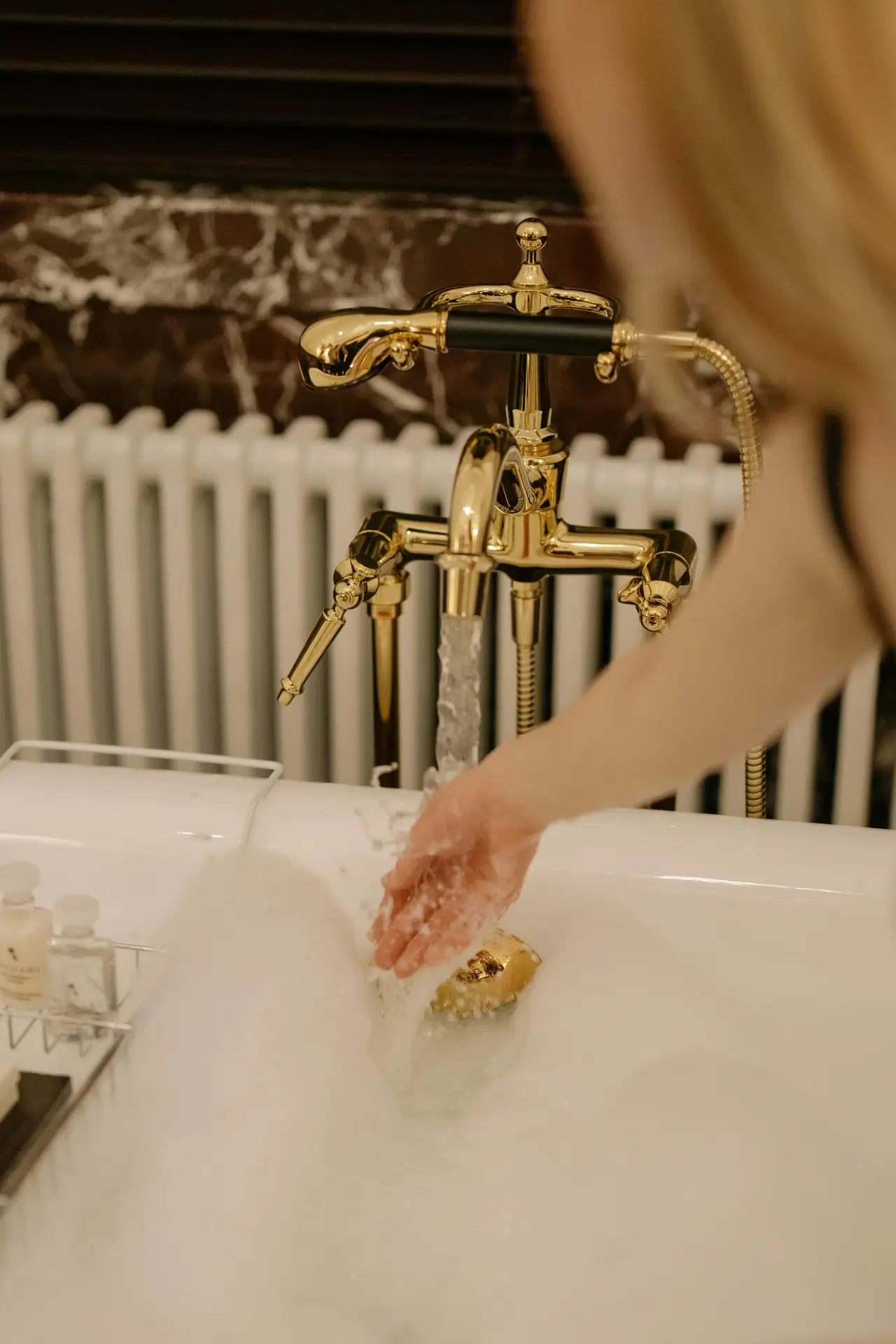 A close-up view of a bathtub filled with soft bubbles, featuring elegant brass fixtures with a handheld shower and flowing water. A hand gently reaches into the bath, adding a relaxed, spa-like feel, while warm tones and classic details highlight girls bathroom ideas that use brass fixtures to bring a touch of luxury and charm.