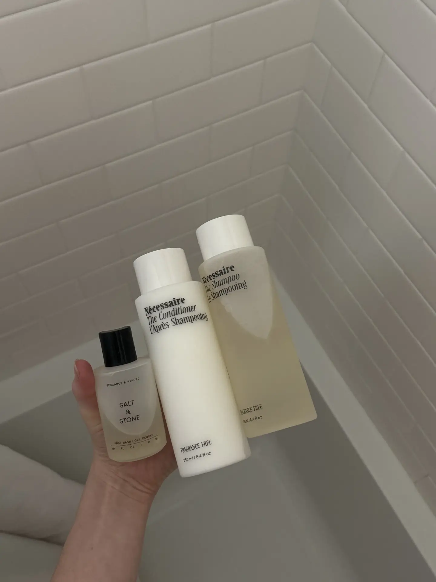 A hand holding three minimalist bath products in matching neutral packaging above a bathtub with white subway tiles. The soft color palette and clean labels showcase girls bathroom ideas that use coordinated bottles to create a calm, cohesive look while keeping everyday shower essentials visually organized.