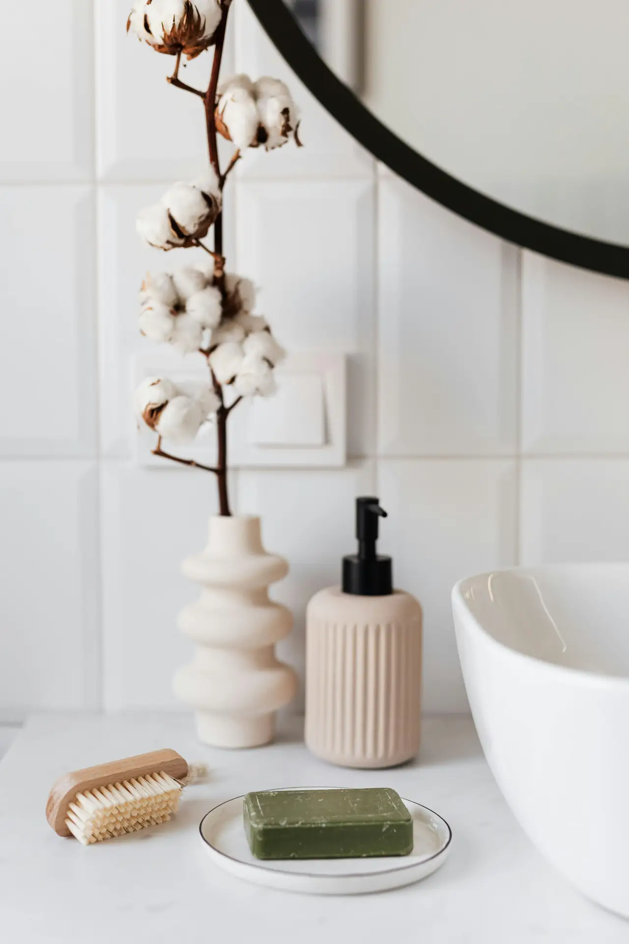 A bright bathroom countertop styled with a ceramic vase holding cotton stems, a ribbed soap dispenser, a small wooden brush, and a decorative soap dish with a green bar of soap beside a modern sink. The setup reflects girls bathroom ideas by combining soft neutrals and simple accessories that work well in many different bathroom styles.