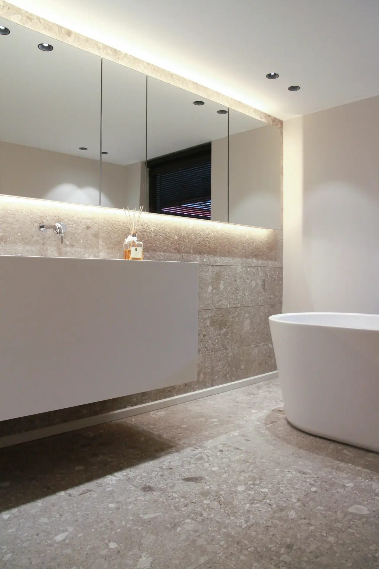 A sleek, contemporary bathroom with a floating vanity, stone-textured walls, and a wide frameless mirror that reflects soft recessed lighting. A freestanding bathtub and neutral finishes create a calm, modern atmosphere, offering girls bathroom ideas that use a frameless mirror to keep the space feeling open and elegant.