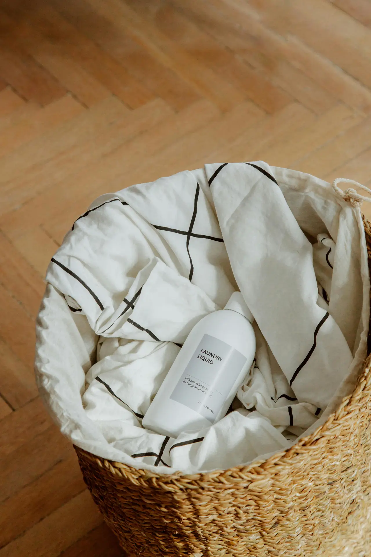 A woven basket placed on a wooden floor, filled with light-colored fabric and a minimal laundry detergent bottle tucked inside. The scene fits girls bathroom ideas by showing a hidden laundry hamper that keeps everyday essentials out of sight while maintaining a clean, natural, and flexible look suitable for different bathroom styles.