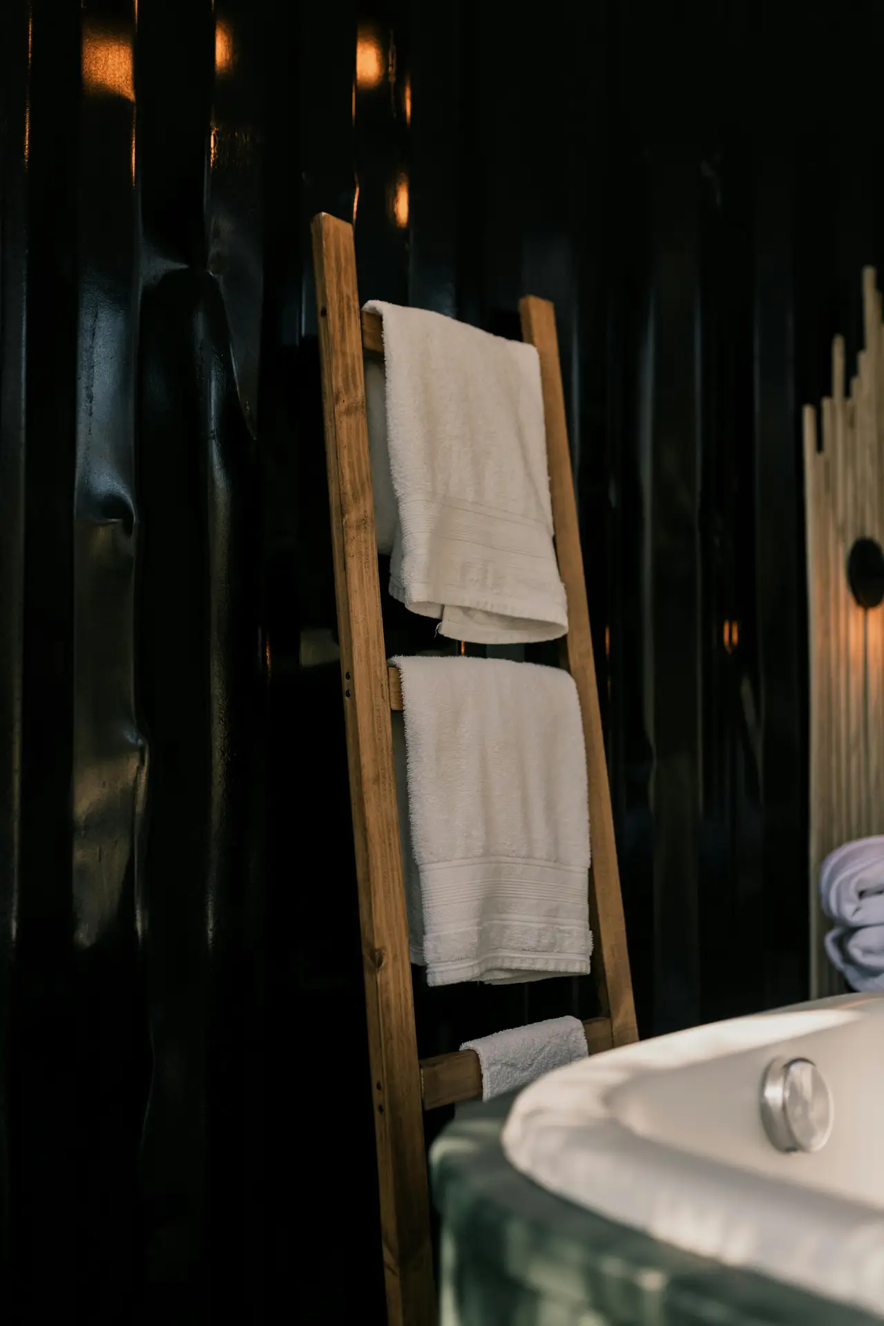 A cozy bathroom corner featuring a wooden ladder rack for towels leaning against a dark, textured wall, with neatly folded white towels adding contrast. Soft lighting and natural materials create a warm, spa-inspired mood, showcasing girls bathroom ideas that use a ladder rack for towels as both a practical storage solution and a decorative accent.