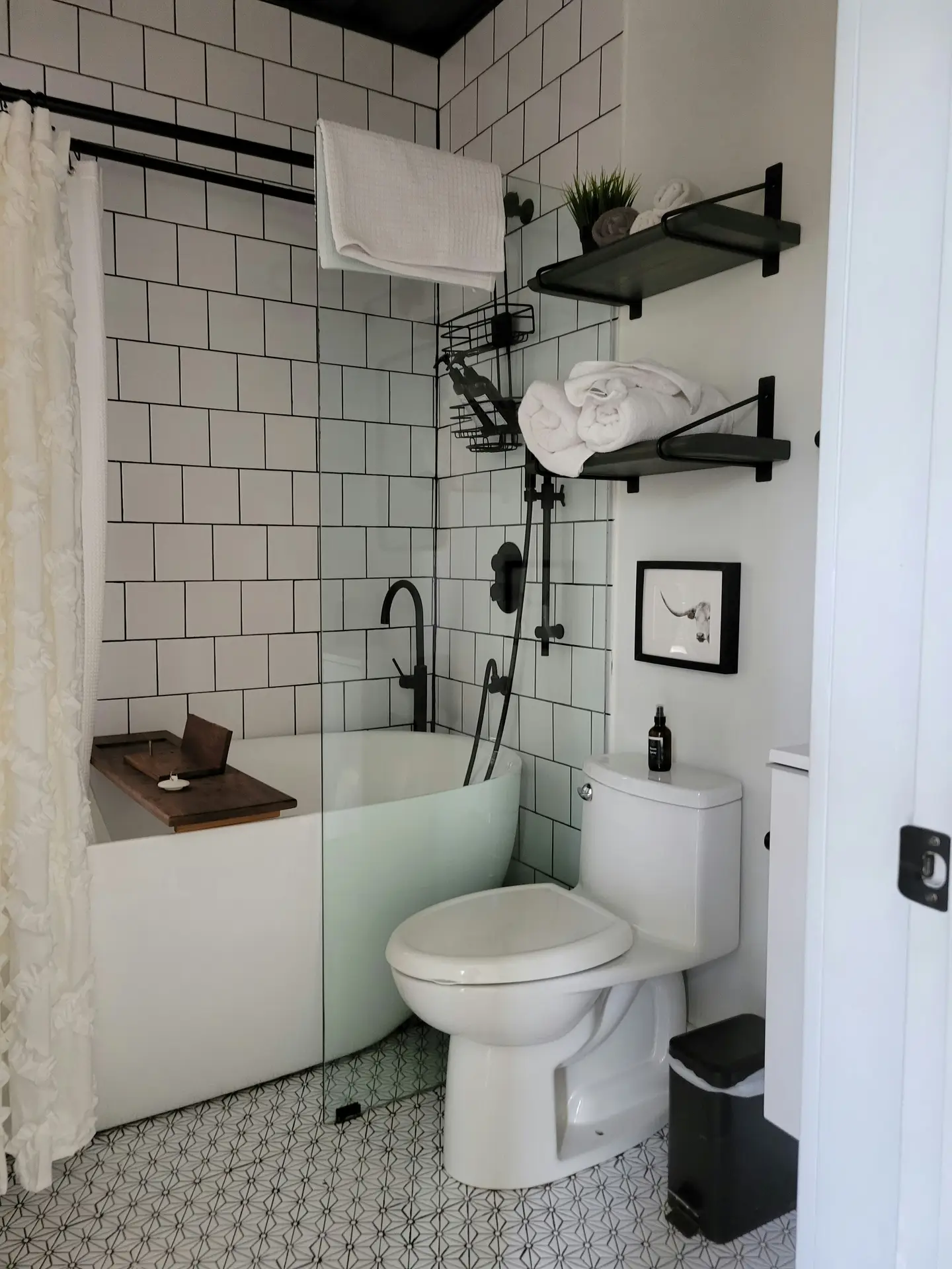 A compact bathroom with white subway tile walls, a glass shower enclosure, and a small soaking tub, featuring black over-toilet shelves styled with rolled towels and simple decor. Matte black fixtures and patterned floor tiles add contrast, presenting girls bathroom ideas that use over-toilet shelves to maximize storage while keeping the space clean and modern.