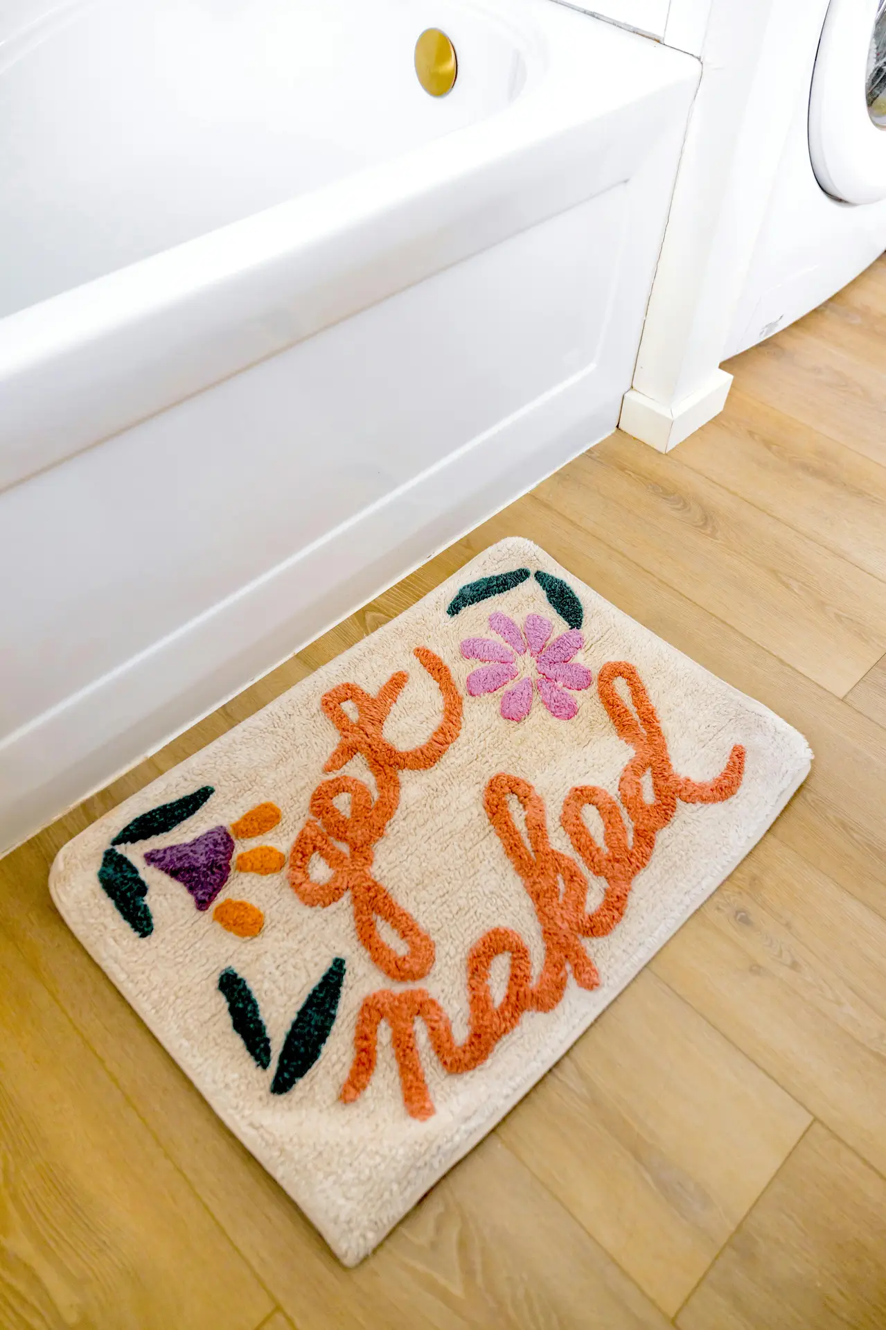 A bright bathroom floor with light wood planks and a white bathtub, styled with a colorful plush rug featuring playful lettering and floral details. The scene fits girls bathroom ideas by showing how a plush rug can add warmth, texture, and a cheerful accent while keeping the space comfortable and easy to personalize.
