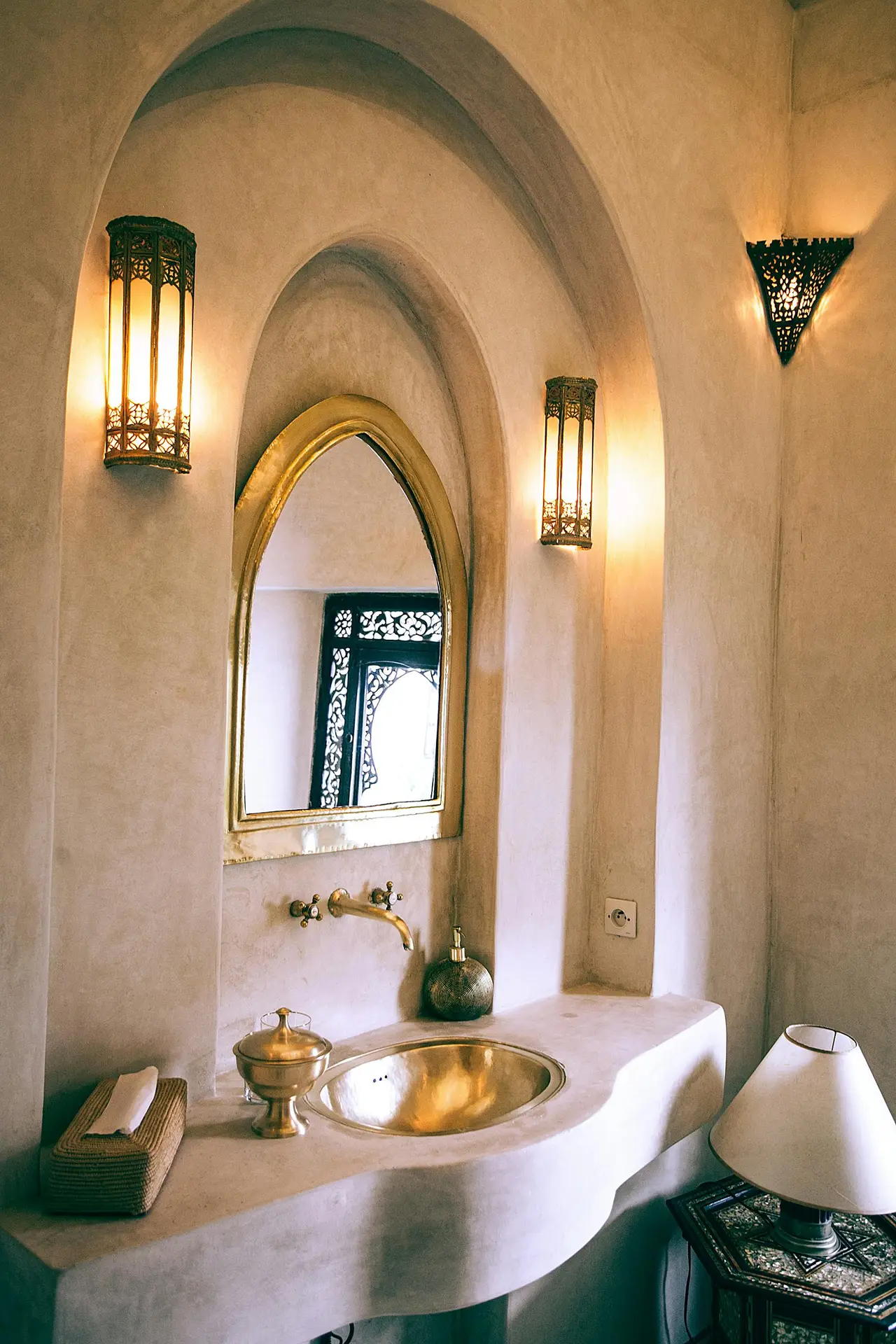 A softly lit bathroom vanity with arched niches, a curved plaster sink, and a brass basin paired with an arched mirror. Warm wall-mounted sconces frame the space, creating an inviting atmosphere that fits girls bathroom ideas inspired by timeless, versatile design with subtle decorative accents.