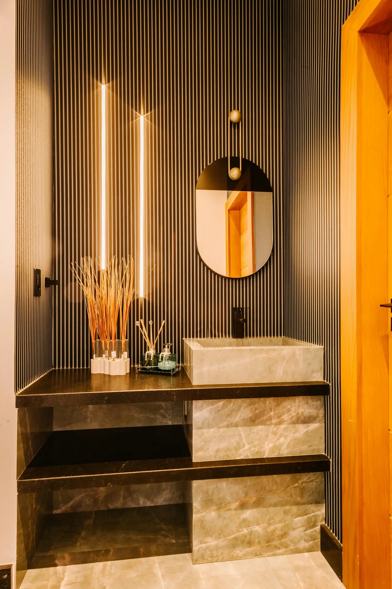 A modern bathroom vanity with a stone sink and open shelving, set against a bold wallpaper accent with vertical lines and warm lighting. This scene reflects girls bathroom ideas by showing how a wallpaper accent can frame the sink area, add depth, and create a stylish focal point that works in both contemporary and cozy spaces.