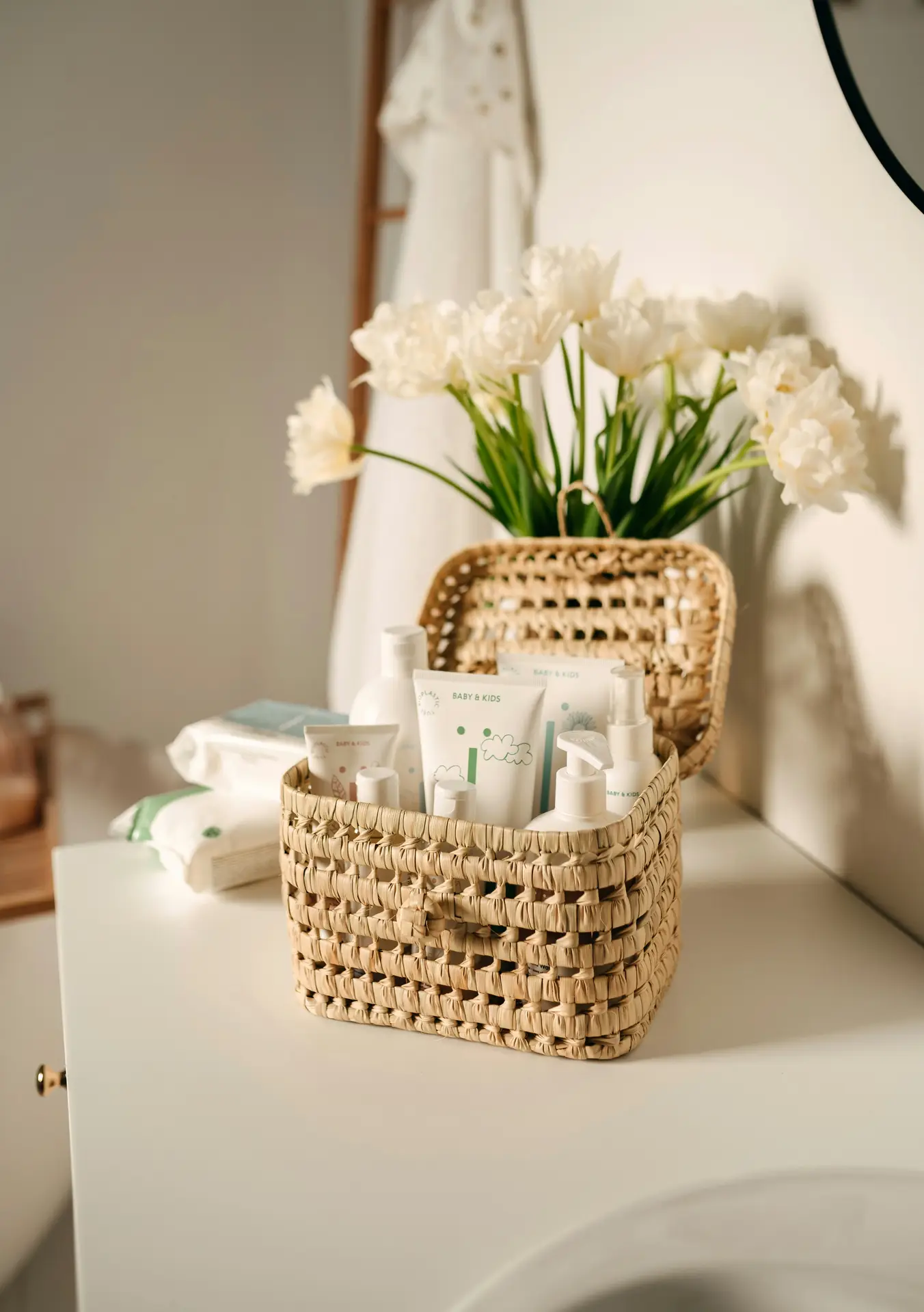 A light, cozy bathroom setup with a woven basket for skincare placed on a white vanity, filled with neatly arranged bottles and wipes. Soft flowers and neutral tones add a calm, welcoming feel, offering girls bathroom ideas that use woven baskets for skin care to keep everyday products organized while adding natural texture to the space.