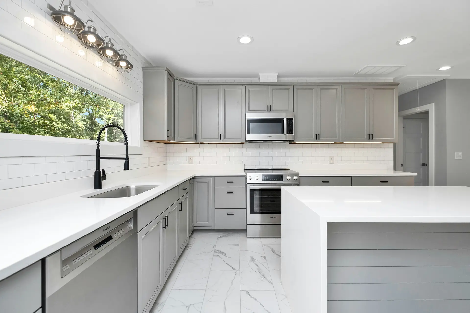 Bright, modern kitchen with gray shaker cabinets, stainless steel appliances, and a large white island; a sleek pull-down faucet and hardware in matte black fixtures add contrast, offering kitchen ideas for a clean and contemporary look.