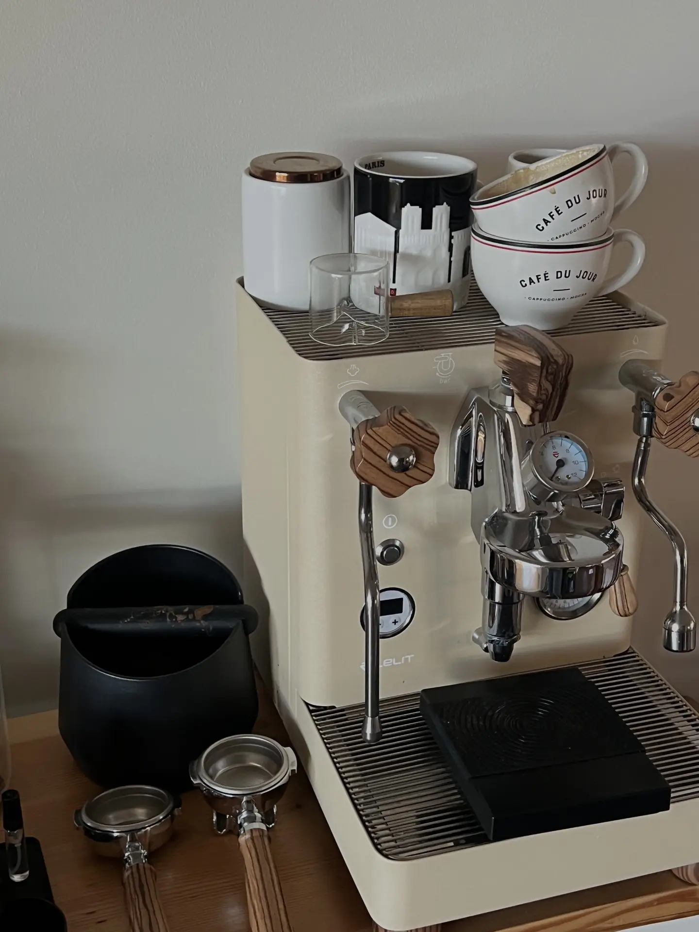 Compact coffee corner featuring a cream-colored espresso machine with wooden accents, mugs and glassware arranged on top, and portafilters and accessories neatly set beside it; a cozy setup that offers kitchen ideas for creating a stylish and functional brewing spot at home.