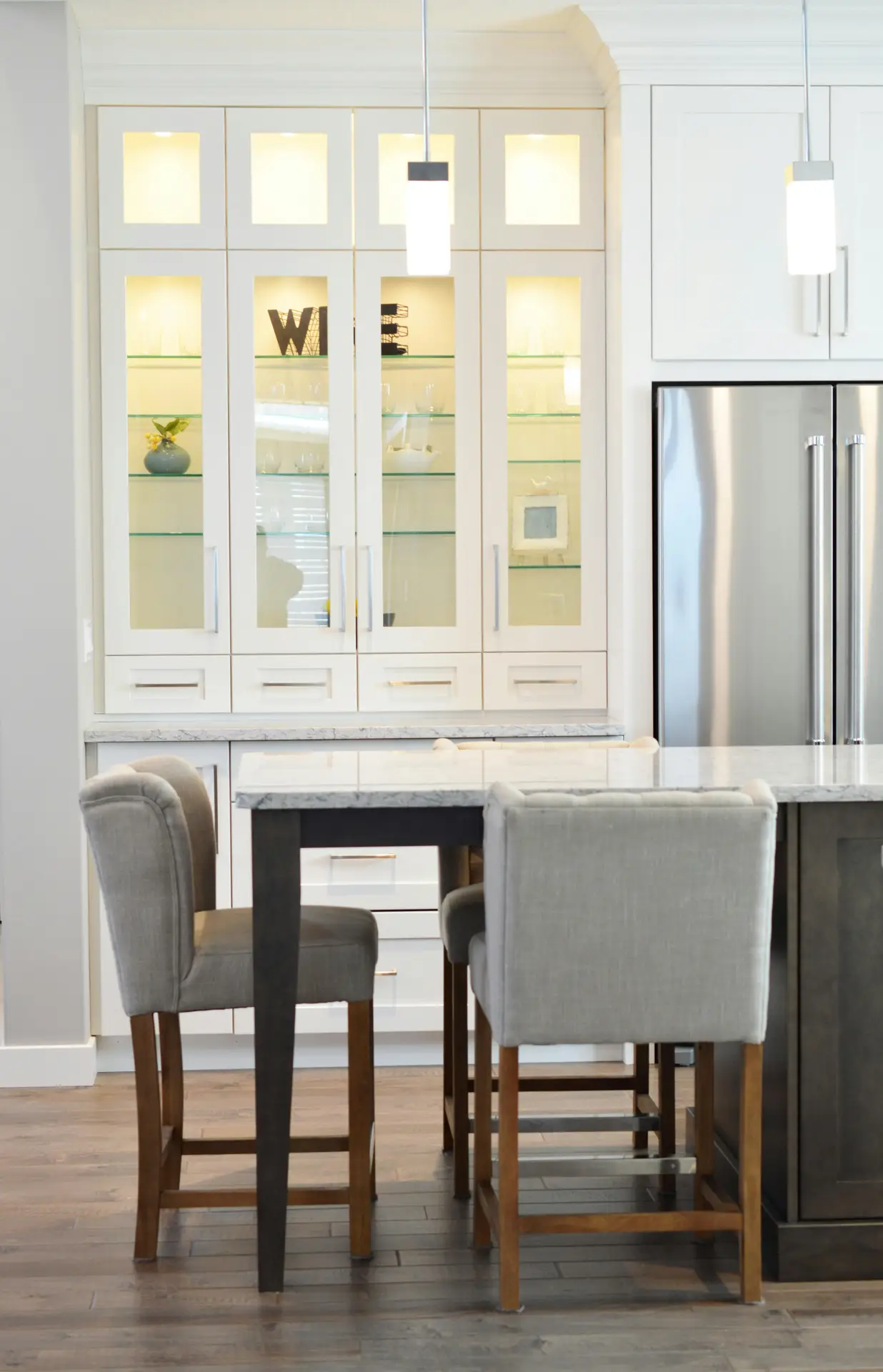 Bright kitchen corner featuring a marble-topped table with upholstered chairs and tall illuminated glass cabinets displaying decor and dishware; the clean design and soft lighting offer kitchen ideas that use glass cabinets to add elegance and openness to the space.