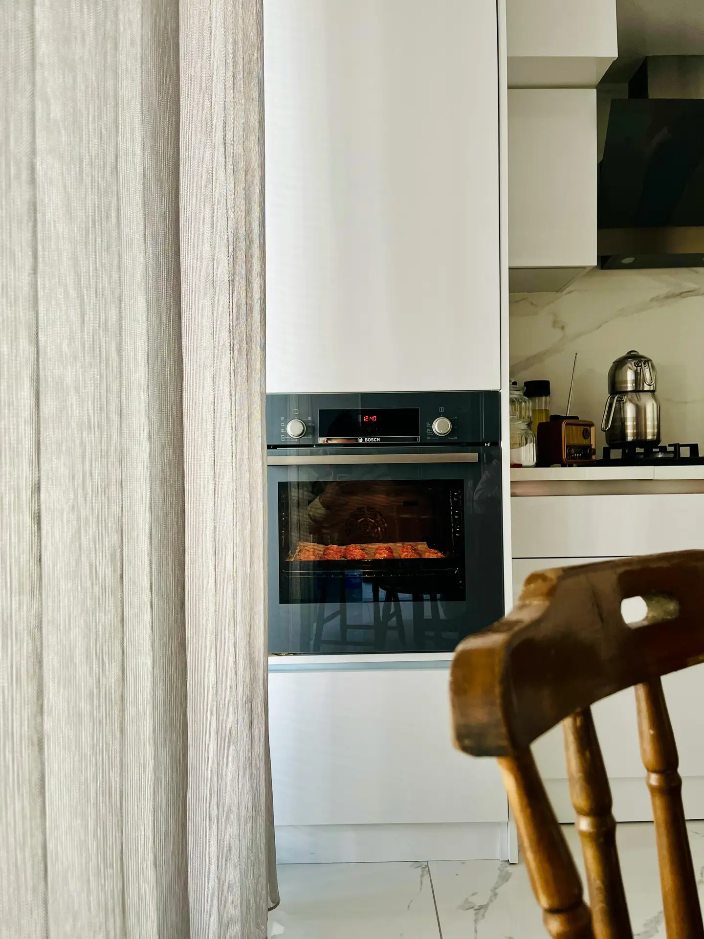 A bright kitchen scene featuring sleek integrated appliances, including a built-in oven set into smooth white cabinetry. Soft neutral curtains filter natural light, while a wooden chair and marble-look flooring add warmth and texture. The setup highlights functional kitchen ideas with a clean, modern aesthetic.