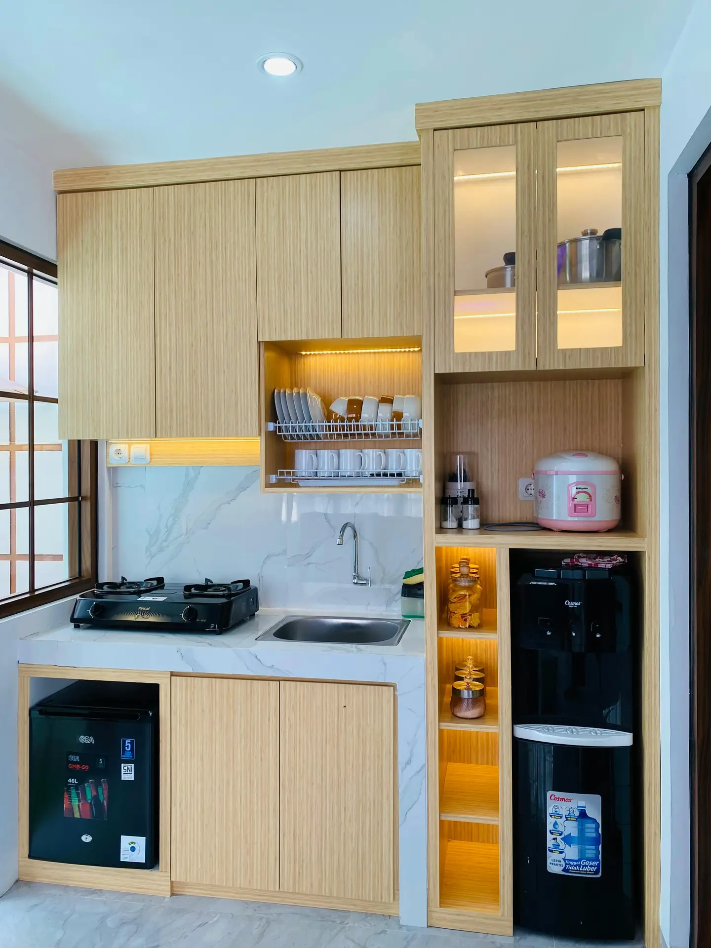 Compact modern kitchen with light wood cabinets, marble-look countertops, open shelving, and warm built-in lighting that highlights layered textures; appliances and dish racks are neatly arranged, offering kitchen ideas for small but efficient spaces.