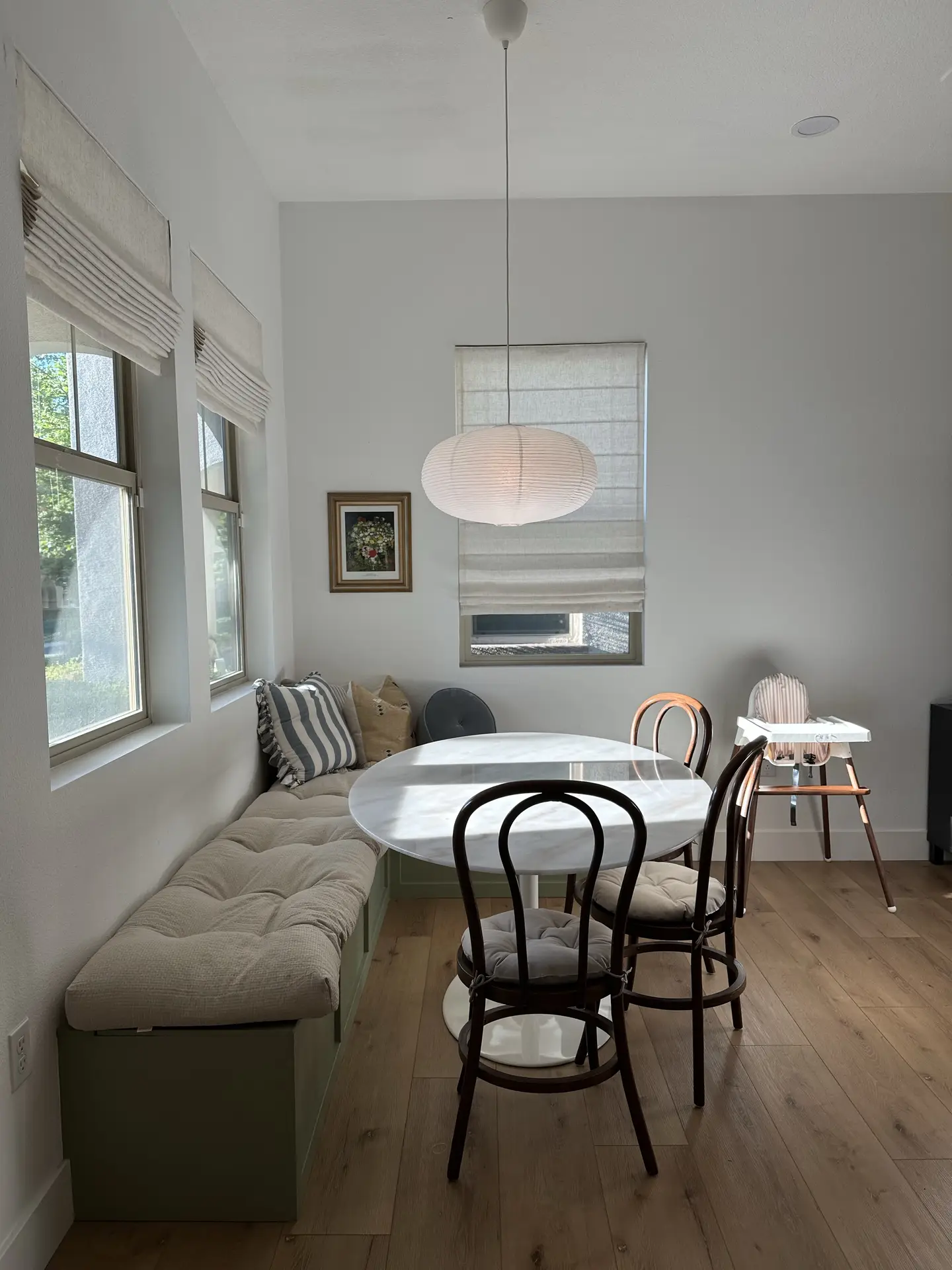Bright dining nook with mixed seating, featuring a built-in cushioned bench, two bentwood chairs, and a round white table; soft neutral tones, layered pillows, and simple window shades create a cozy look, offering kitchen ideas for small, inviting breakfast spaces.