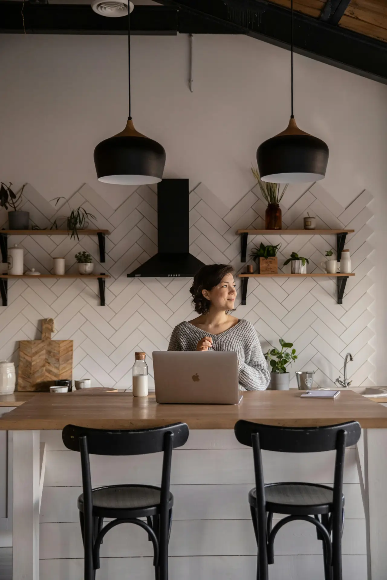 Cozy kitchen scene with a woman working at a wooden island, surrounded by open shelves holding plants and ceramics, black pendant lights, and a herringbone tile backsplash; the mix of wood, greenery, and simple finishes highlights natural materials and offers kitchen ideas for warm, lived-in spaces.