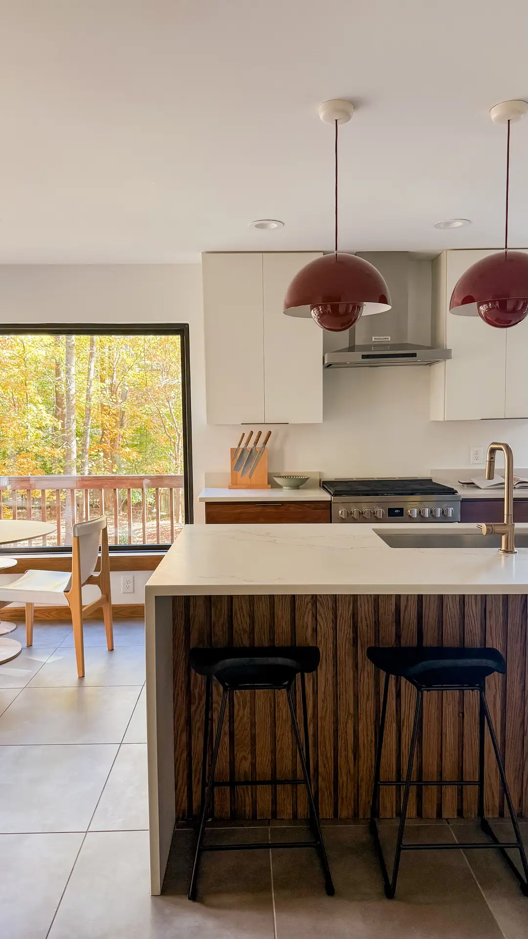 Modern kitchen with a wood-accent island, sleek cabinetry, and brass fixtures, illuminated by two oversized burgundy statement pendant lights; large windows frame the outdoor view, offering kitchen ideas that blend warm tones with contemporary design.