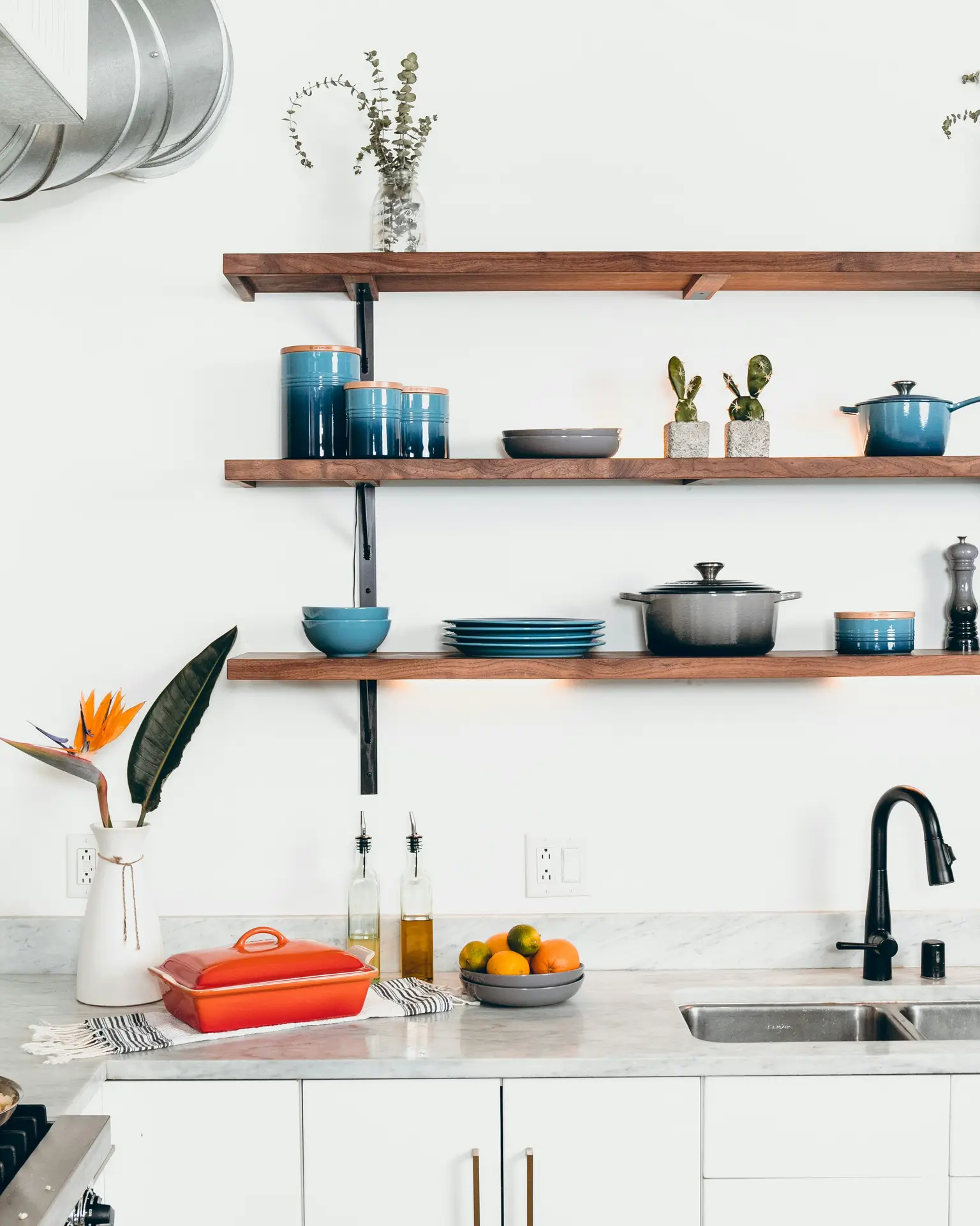Modern kitchen with marble countertops, open wood shelving, and a mix of colorful dishes and cookware, accented by fresh greenery and bold accessories; the thoughtful styling highlights signature details and offers kitchen ideas for creating a curated, welcoming space.