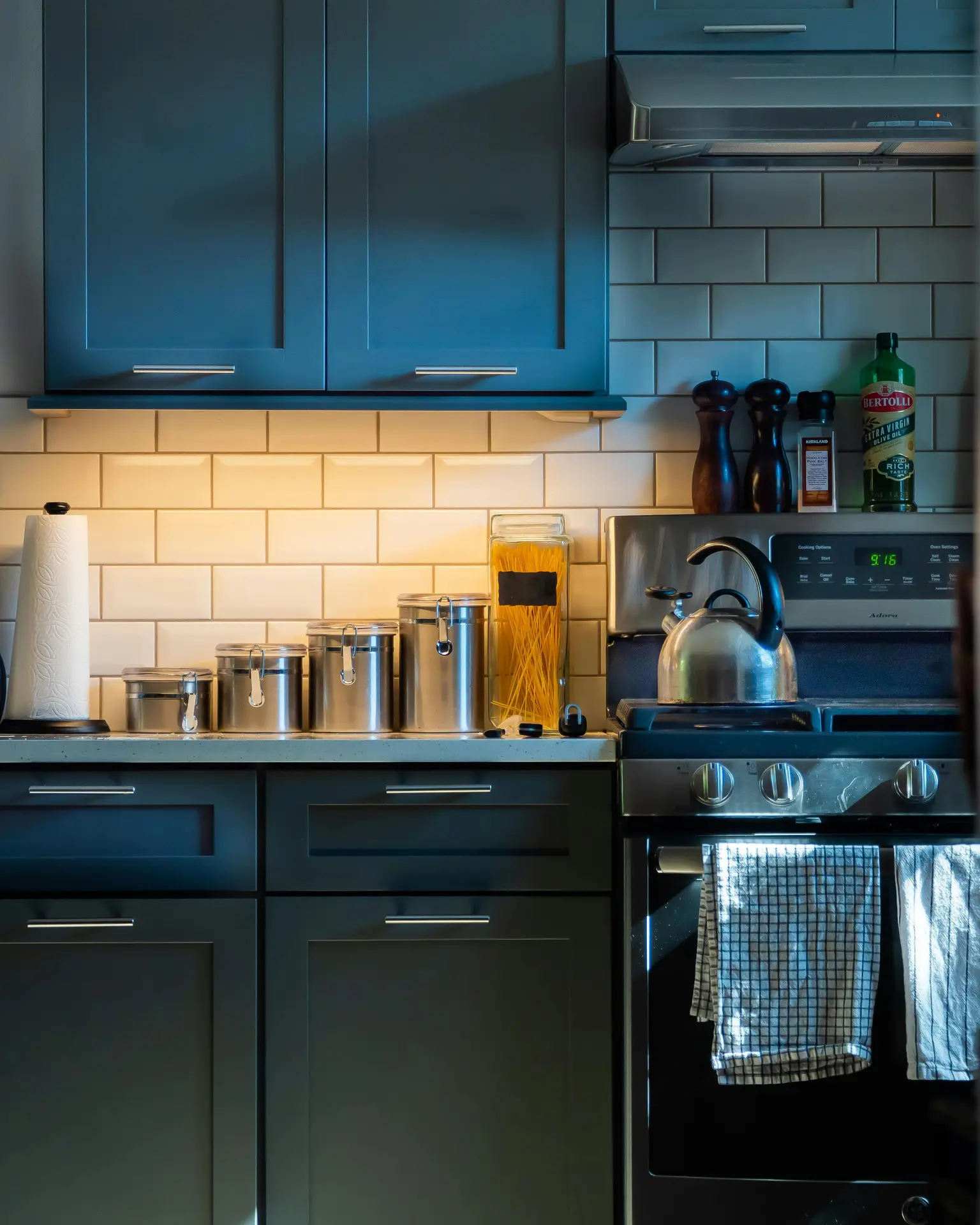 Cozy kitchen setup with deep blue cabinets, stainless steel canisters, and a stove with tea towels, set against a warm, textured backsplash of glossy subway tiles; the mix of colors and lighting offers kitchen ideas for creating a stylish and practical cooking space.