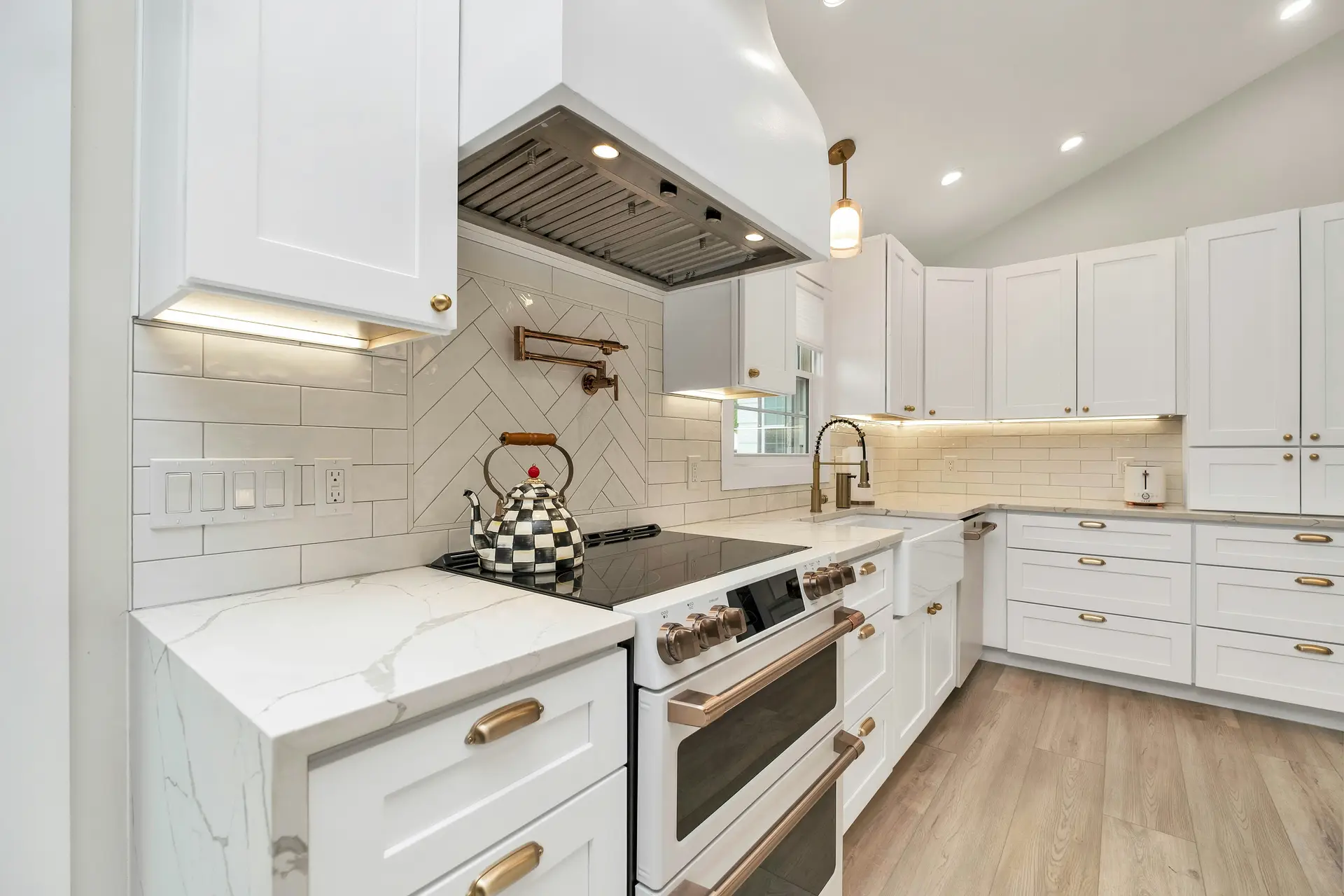 Bright white kitchen with shaker cabinets, marble-look countertops, and mixed tile patterns around the stove; warm under cabinet light highlights the brass hardware and fixtures, offering kitchen ideas that blend classic style with modern function.