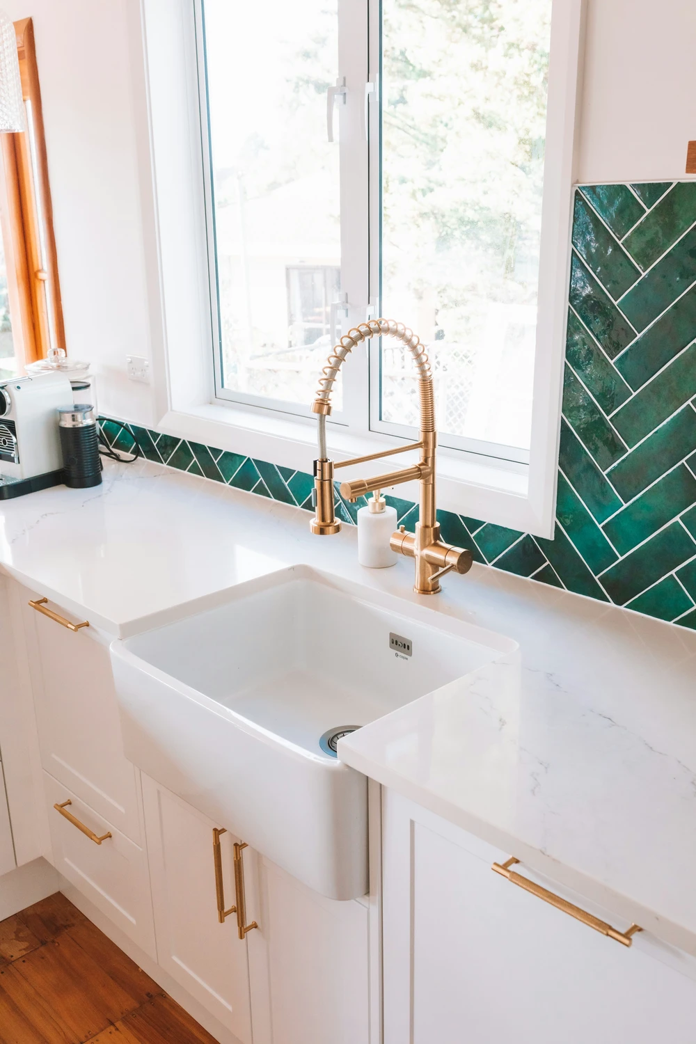 Bright kitchen sink area with a farmhouse basin, white countertops, and green herringbone tile backsplash; a brass pull-down faucet and matching hardware add warm metallic touches, offering kitchen ideas that blend fresh color with elegant detailing.