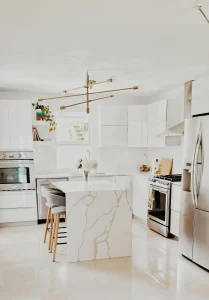 Bright, modern kitchen with glossy white cabinets, stainless-steel appliances, and a marble waterfall countertop island with bar stools; a gold linear chandelier and minimal decor add a clean, stylish touch, perfect for kitchen ideas focused on airy, contemporary spaces.