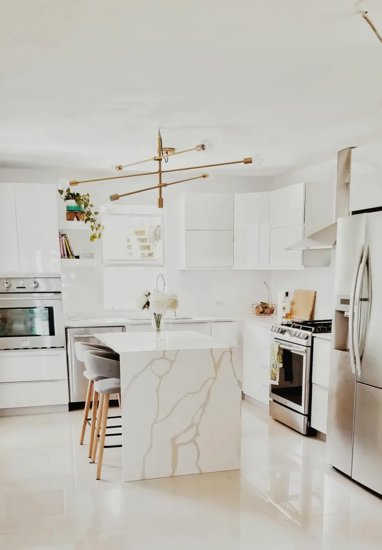 Bright, modern kitchen with glossy white cabinets, stainless-steel appliances, and a marble waterfall countertop island with bar stools; a gold linear chandelier and minimal decor add a clean, stylish touch, perfect for kitchen ideas focused on airy, contemporary spaces.