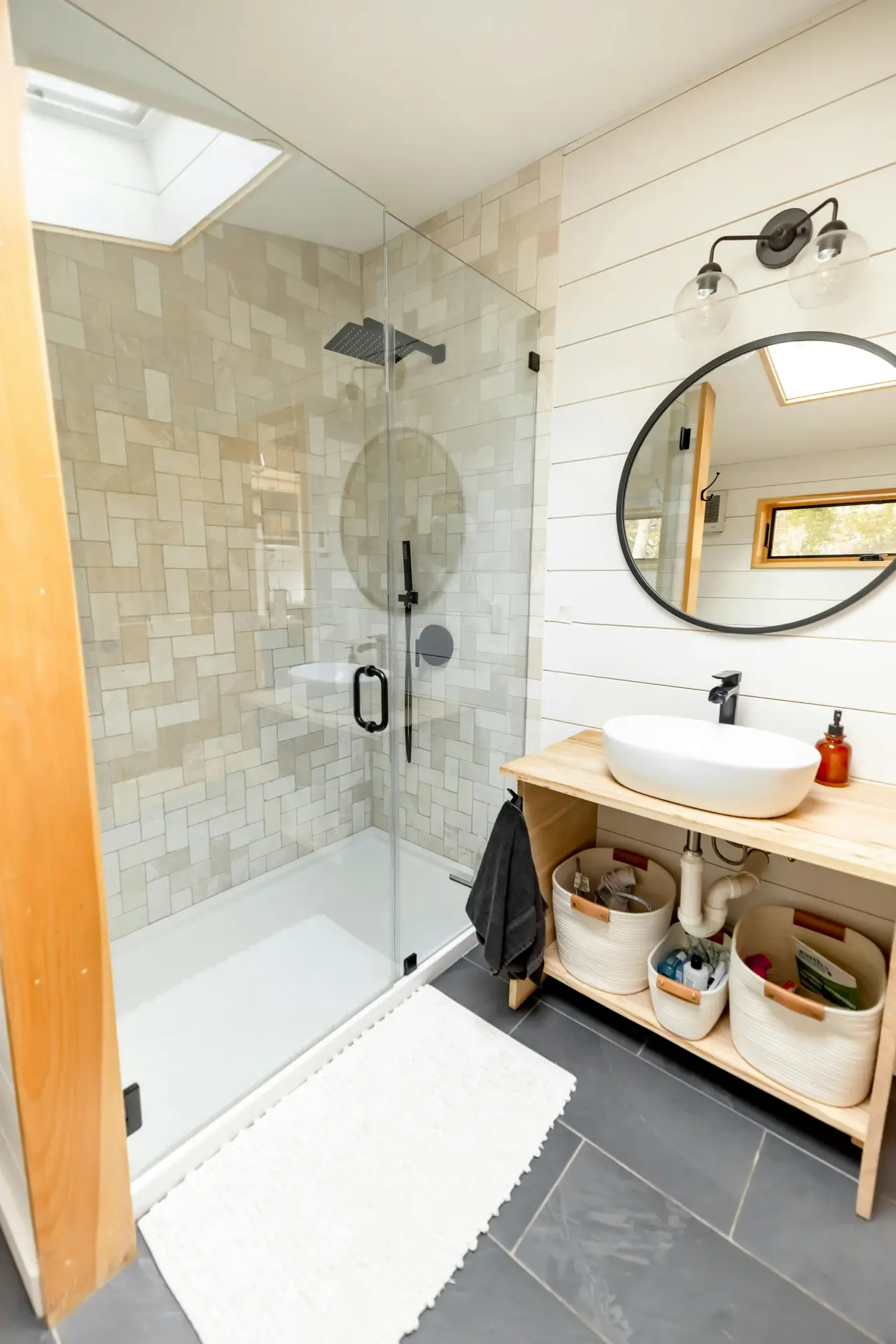 A bright, modern bathroom featuring a glass shower enclosure with an accent niche framed in contrasting tiles that add subtle depth to the wall. A round mirror hangs above a wooden vanity with a vessel sink, while woven baskets provide open storage beneath. Soft lighting and clean lines offer small bathroom decor ideas that highlight how an accent niche with contrasting tiles can elevate even a compact space.