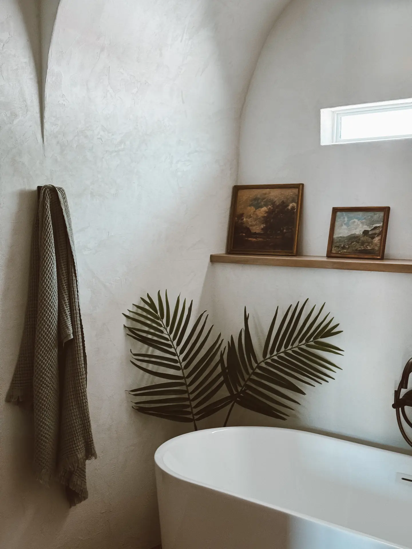 A serene bathroom with textured plaster walls and soft, natural lighting, featuring a freestanding tub beneath a slim ledge that holds two vintage-style art prints. A palm leaf arrangement and a textured towel add organic detail, creating a calm, curated look. The mix of artwork and greenery offers small bathroom decor ideas that bring personality and warmth through simple art prints and natural accents.