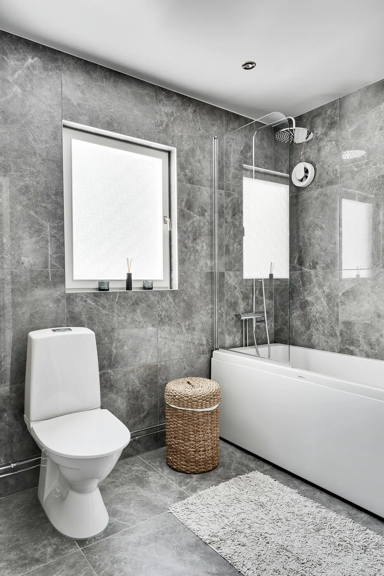 A sleek, modern bathroom with gray marble-look tiles, a white bathtub with a glass shower screen, and a simple toilet. A woven hamper and a soft textured bath mat add warmth and contrast to the cool tones, while a small reed diffuser sits on the window ledge. The clean layout and subtle accents offer small bathroom decor ideas that use a bath mat and natural materials to create a cozy, balanced feel.