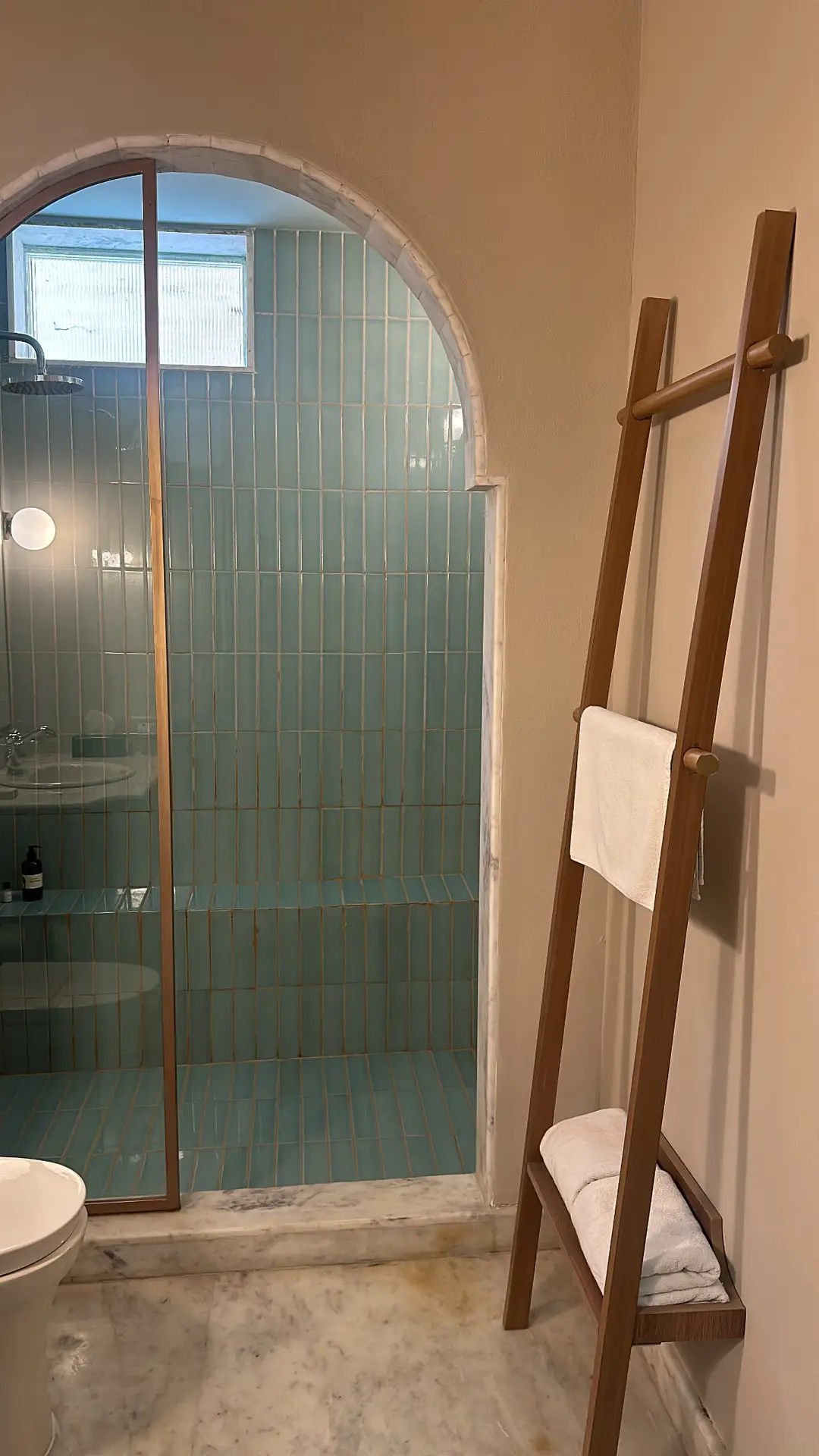 A cozy bathroom with soft beige walls and a marble floor, featuring a walk-in shower framed by an arched opening and lined with vertical teal tiles. A wooden ladder rack stands nearby, holding neatly folded towels and adding a warm, functional accent. The mix of natural materials, subtle color contrasts, and simple storage solutions offers fresh inspiration for small bathroom decor ideas that balance charm and practicality.
