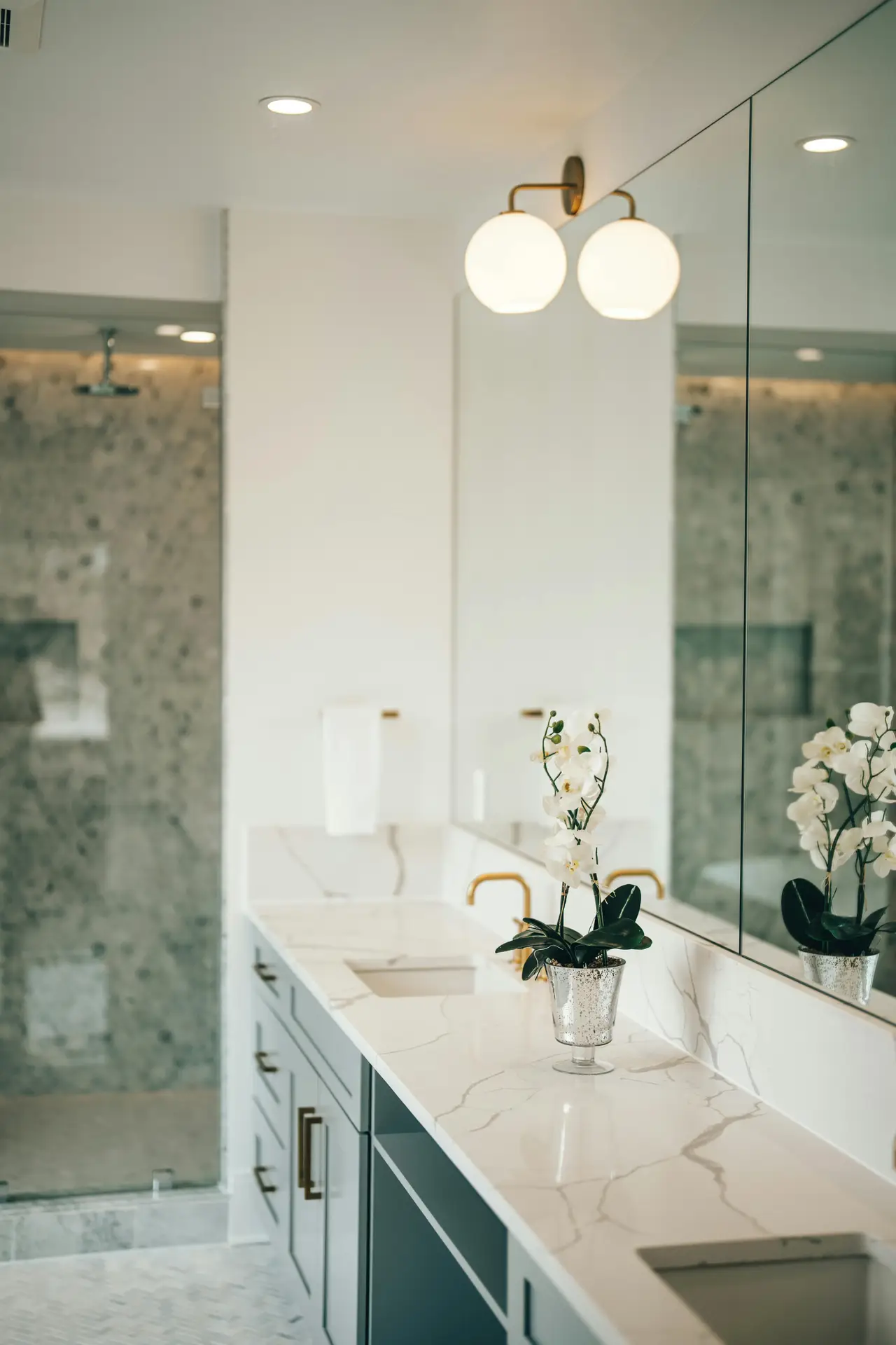 A bright, modern bathroom with a long marble countertop, gold fixtures, and a large mirror that reflects soft overhead lights and a pair of globe sconces, creating a layered lighting effect. A small potted orchid adds a touch of elegance beside the sink, while the glass shower in the background keeps the space open. The clean palette and thoughtful illumination offer small bathroom decor ideas that use layered lighting to elevate both style and function.