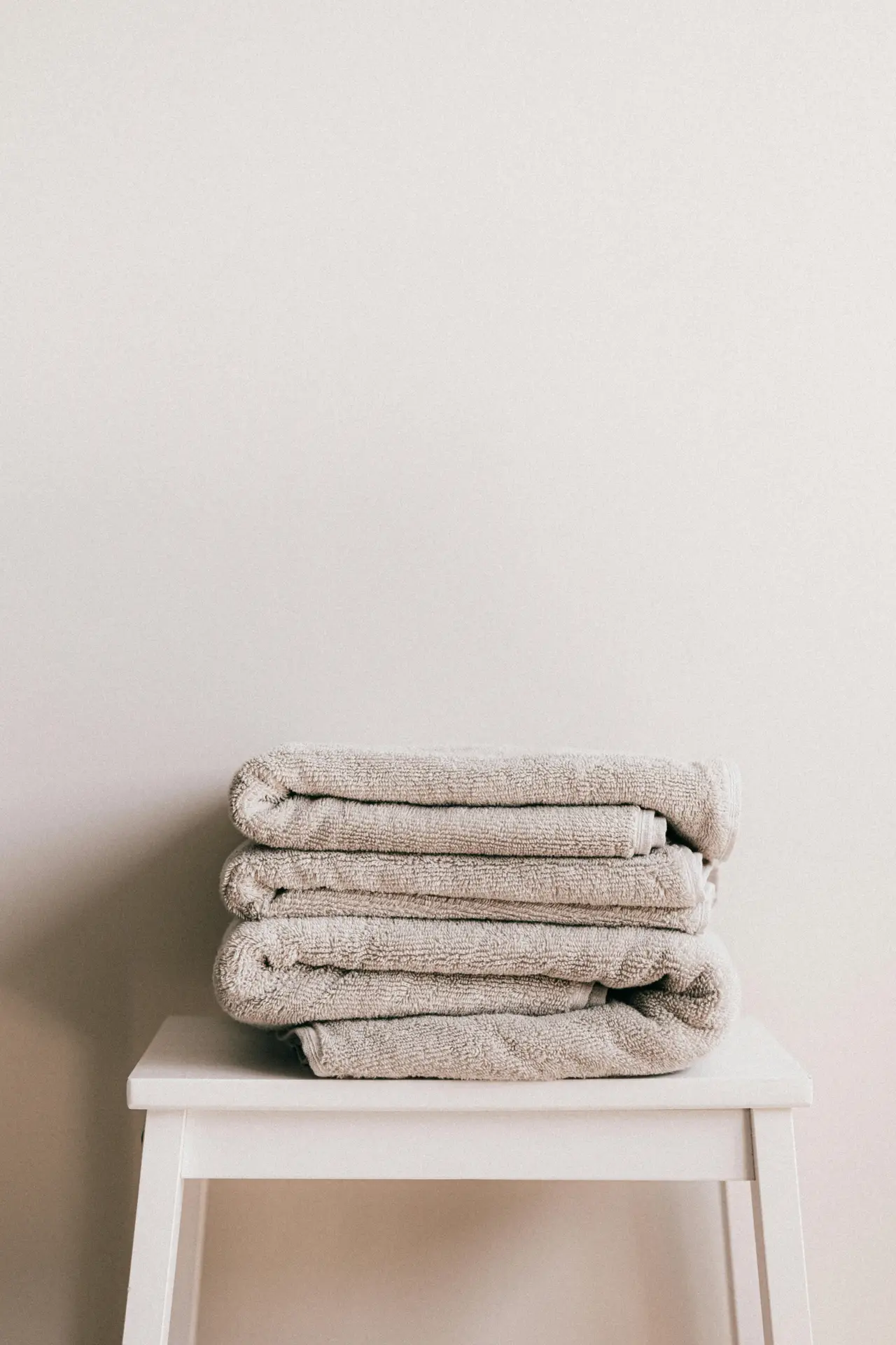 A neatly folded stack of soft textured towels resting on a simple white stool against a neutral wall. The clean, minimal setup highlights how small bathroom decor ideas can feel cozy and refined by incorporating plush towels as both a practical essential and a subtle decorative touch.