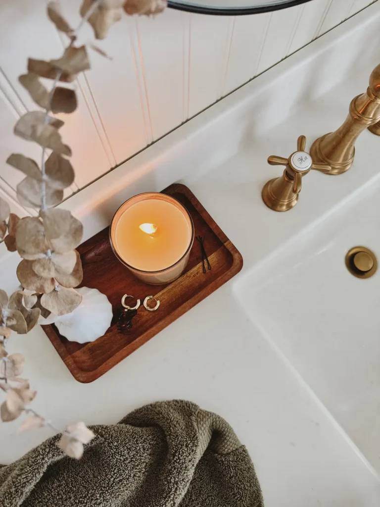 A cozy bathroom countertop styled with a wooden tray holding a glowing spa-like candle, a small vase, hair pins, and gold earrings. Nearby, soft towels and brass faucet fixtures add warmth and charm. The inviting setup offers small bathroom decor ideas that use spa-like candles to create a relaxing, elevated atmosphere even in compact spaces.