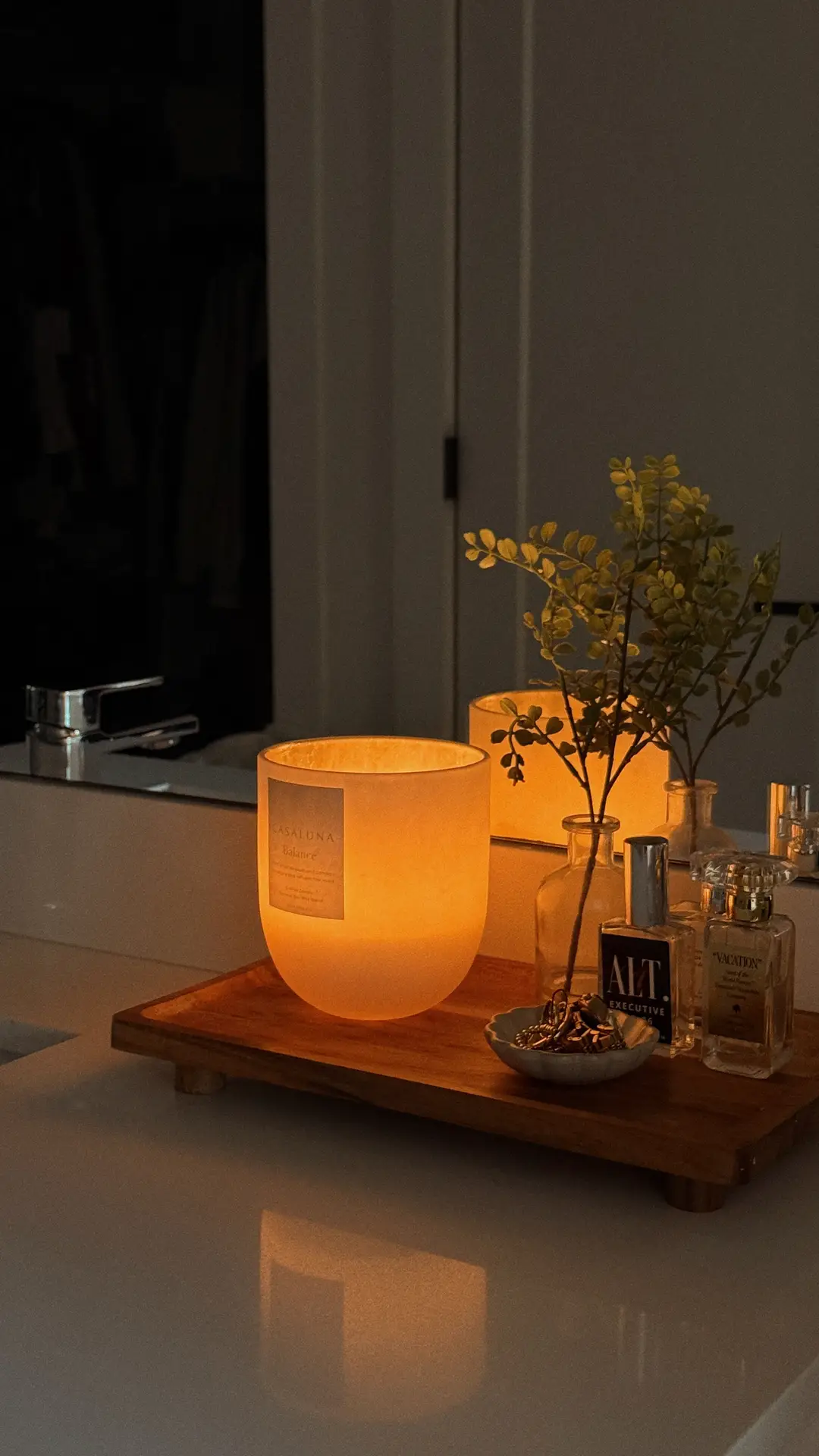 A softly lit bathroom vanity styled with a glowing candle placed on a wooden tray, surrounded by small perfume bottles and a simple glass vase with greenery. The setup reflects girls bathroom ideas by creating a calm candle corner that adds warmth, ambiance, and an inviting spa-like feel without overpowering the space.