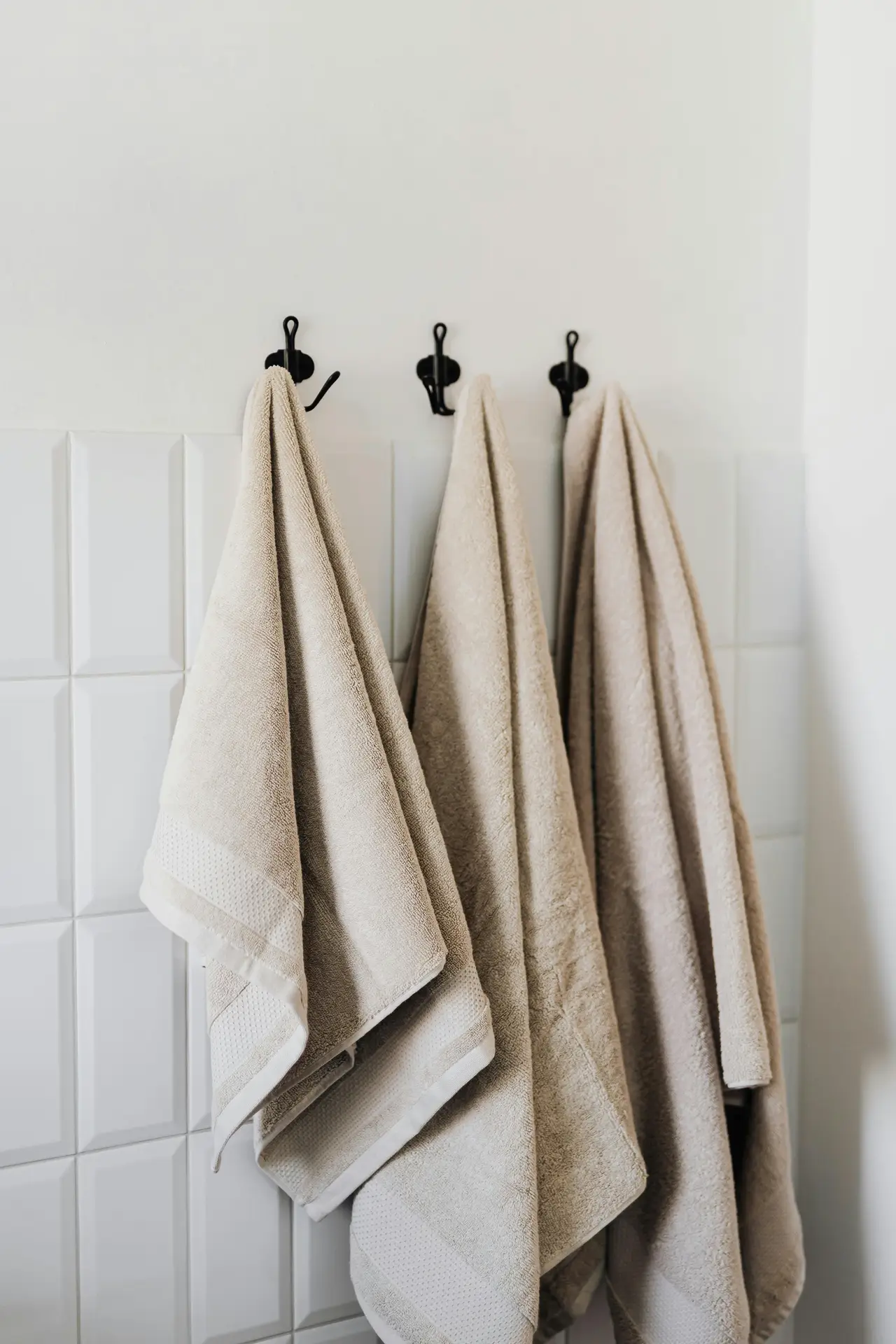 A set of neutral towels hanging neatly from simple black wall hooks mounted above white textured tiles. The soft tones and practical layout create an organized, inviting look, offering small bathroom decor ideas that make use of wall hooks to save space while keeping everyday essentials easy to reach.