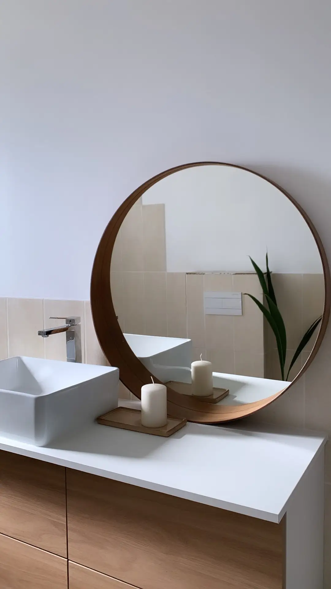 A bright, minimalist bathroom with a round mirror framed in warm wood accents, paired with a sleek white countertop and a modern vessel sink. Two simple candles rest on a wooden tray, while a tall plant adds a natural touch in the reflection. The calm palette and subtle details offer small bathroom decor ideas that use wood accents to bring softness and balance to a clean, contemporary space.