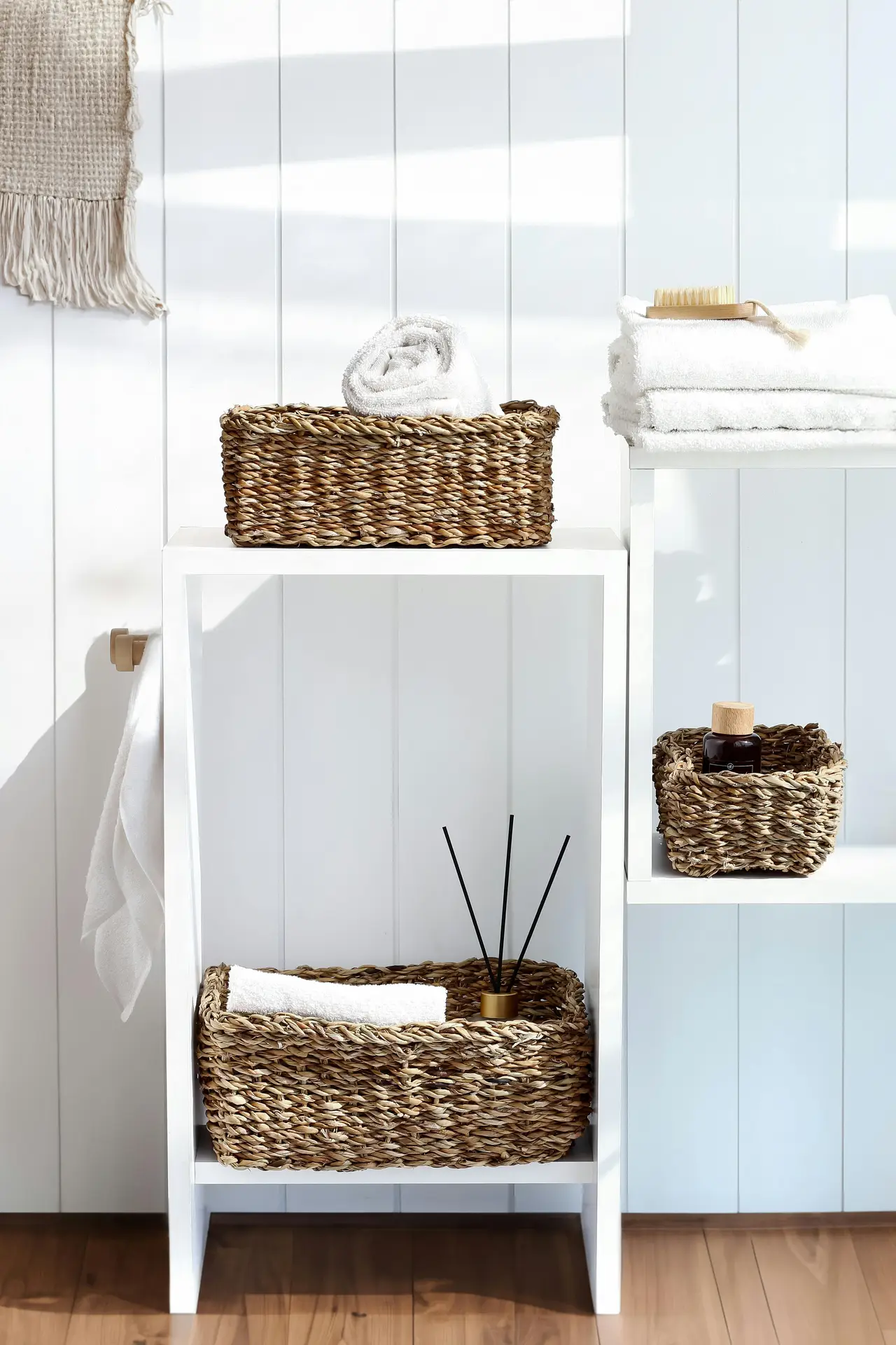 A bright, airy bathroom corner with white shelving styled with woven baskets holding towels, toiletries, and a reed diffuser. The natural texture of the baskets adds warmth against the crisp backdrop, creating a clean yet inviting setup. This arrangement offers small bathroom decor ideas that use woven baskets for practical storage while keeping the space visually calm and organized.