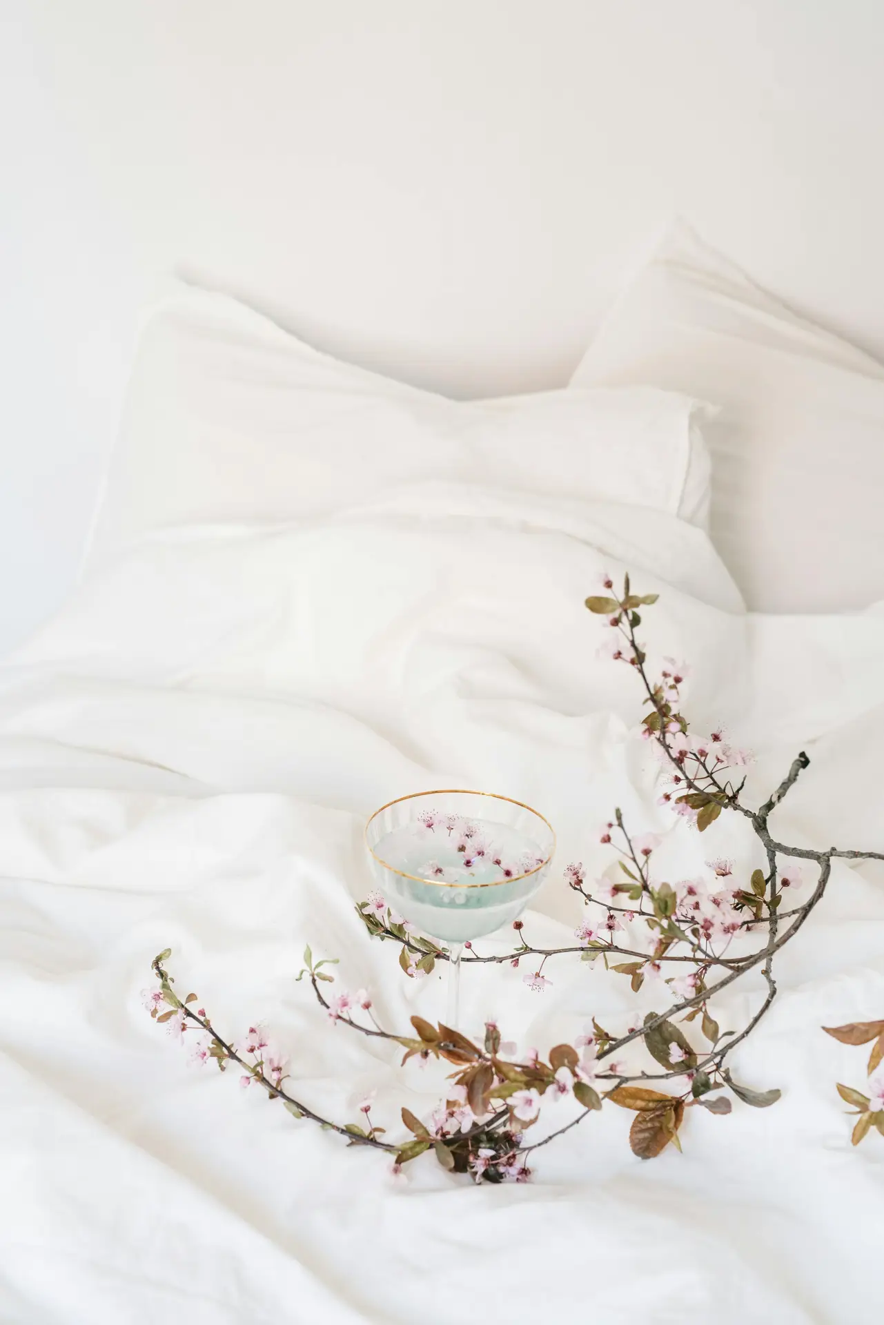 Soft spring decor ideas styled on crisp white sheets, featuring an airy bed layered with light pillows, delicate flowering branches, and a glass filled with petals, creating a calm, fresh setting that feels minimal, seasonal, and adaptable to different bedroom styles.