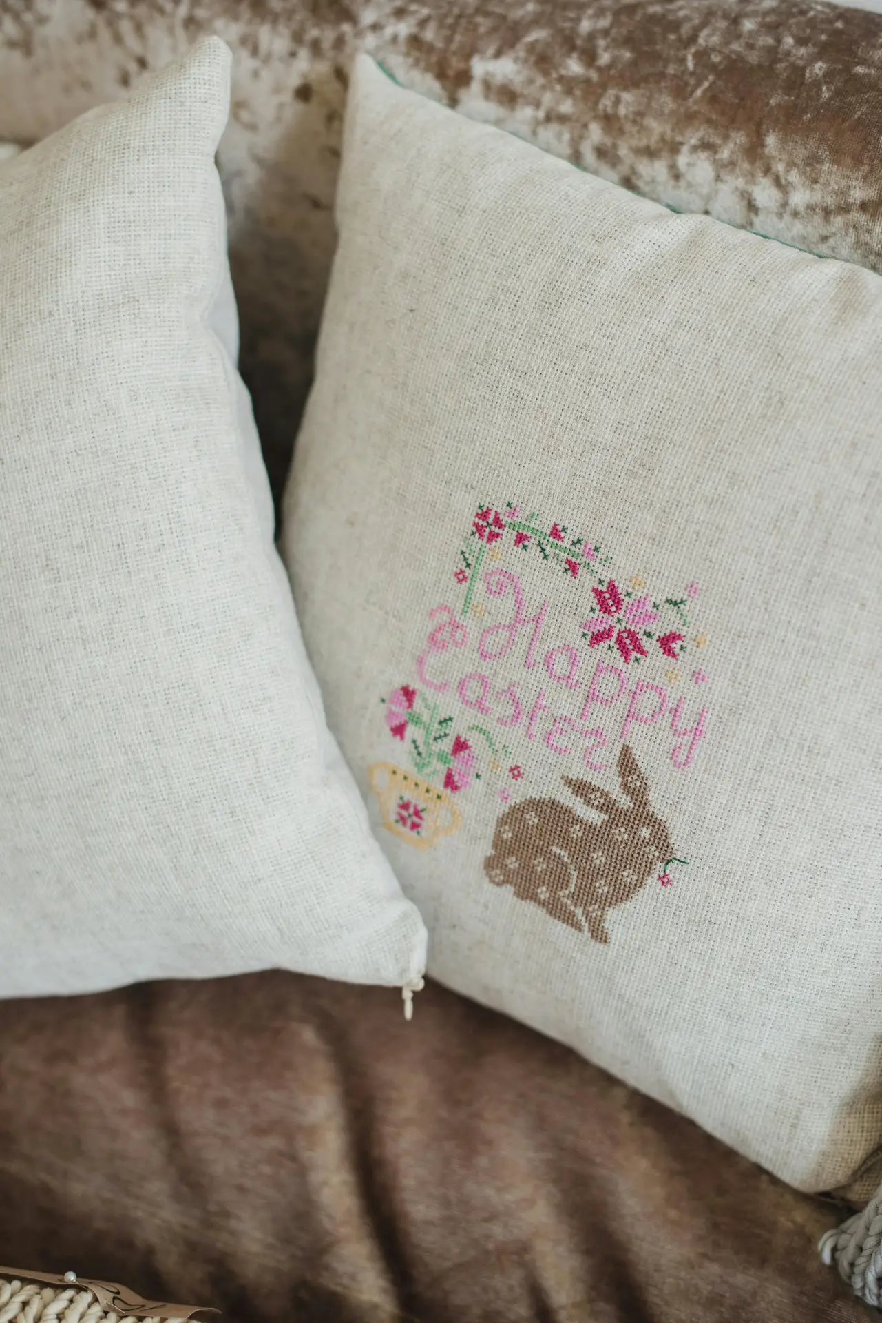 Neutral linen pillows resting on a soft sofa, featuring delicate embroidery with flowers, a small bunny motif, and pastel lettering. The subtle stitching and muted colors add gentle spring decor ideas with charming easter details that work well in cozy living rooms, bedrooms, or seasonal lounge corners.