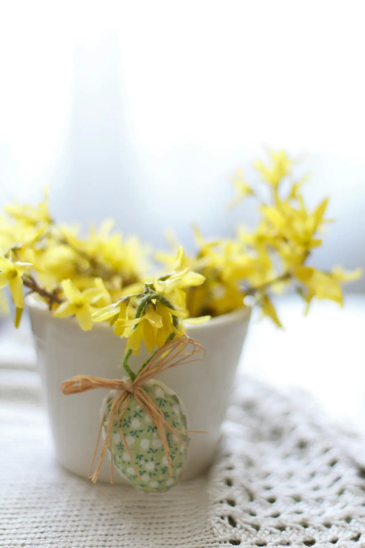 Spring decor ideas showing a small white pot filled with bright yellow forsythia branches, accented by a decorative forsythia with easter egg tied with twine, placed on a light textile surface for a soft seasonal table or windowsill arrangement.