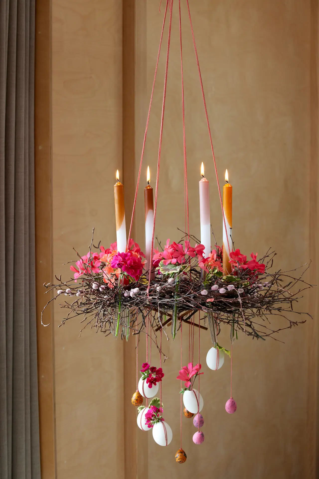 A hanging spring wreath made from a natural twig base is suspended from the ceiling with pink strings, decorated with bright pink flowers, small branches, and dangling pastel eggs. Taper candles glow above the arrangement, creating an airy focal point that fits spring decor ideas and brings a festive, handcrafted look to the room.