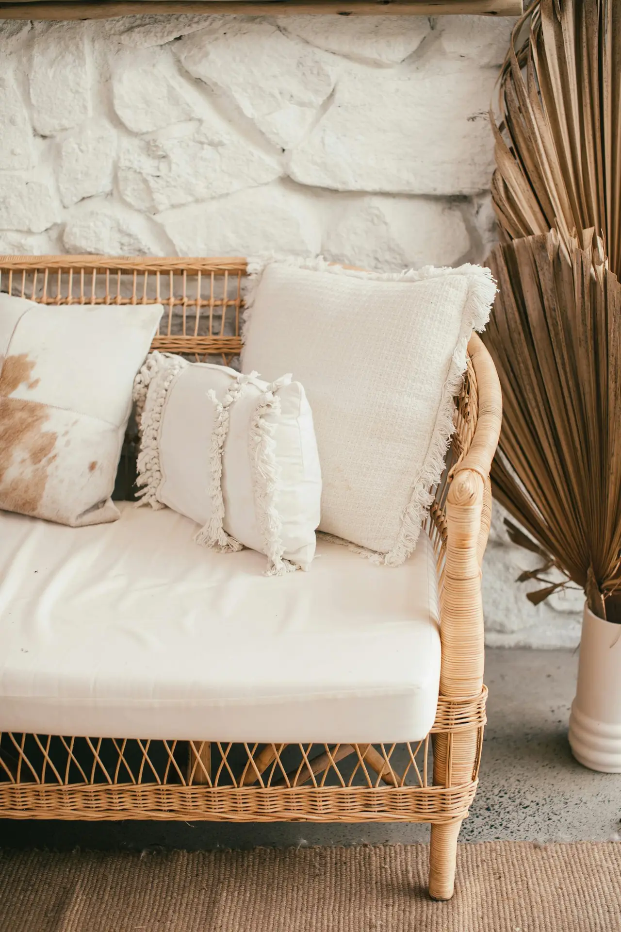 Spring decor ideas shown through a cozy wicker bench styled with light-colored cushions in soft neutral tones, set against a textured stone wall and paired with natural woven accents for a calm, airy seating area.