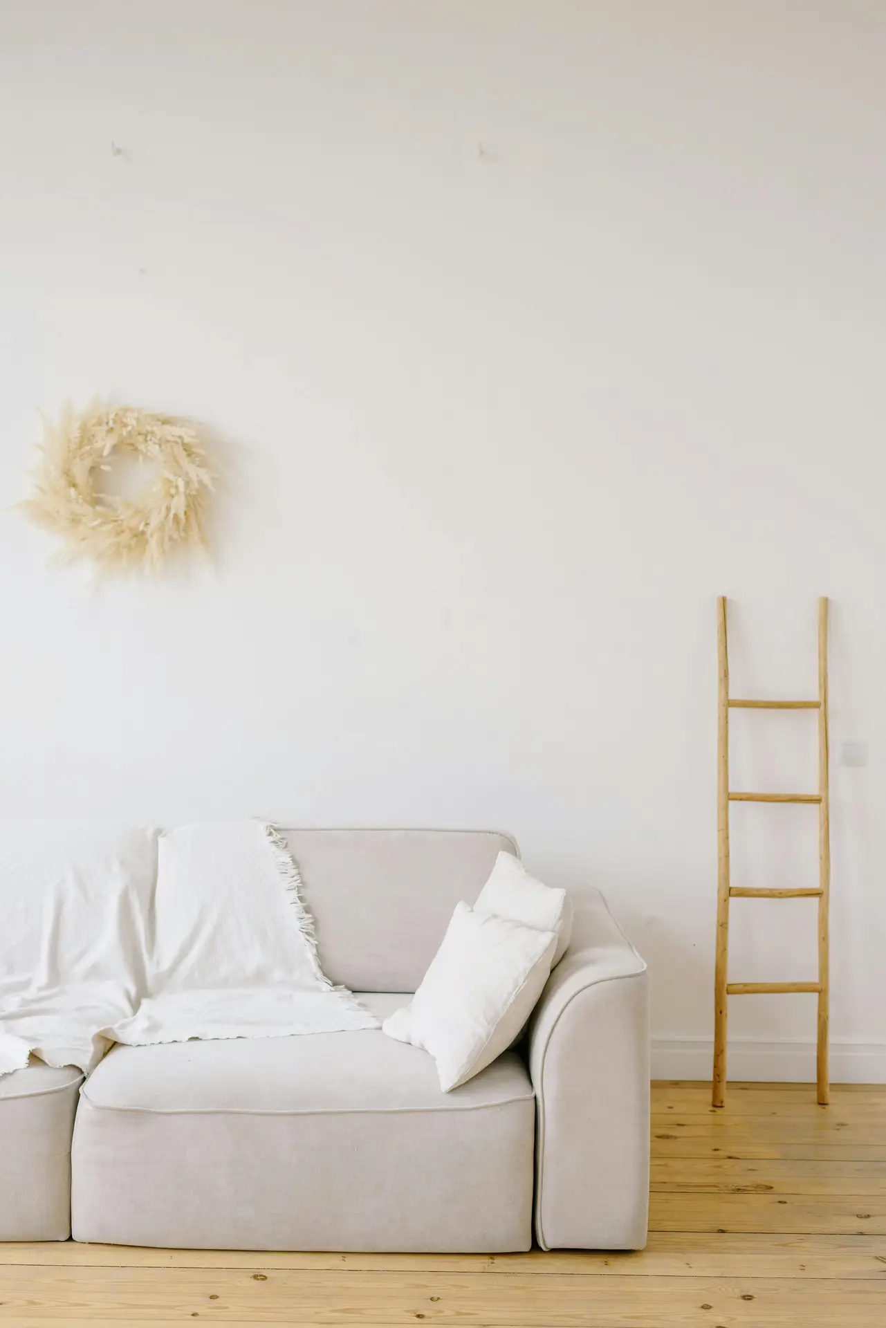 A bright, minimalist living space with a light neutral sofa styled with soft cushions and a linen blanket, set against a white wall and warm wooden floor. A simple wooden ladder and subtle wall decor add texture, making this scene a calm example of spring decor ideas that feel airy, relaxed, and easy to adapt.