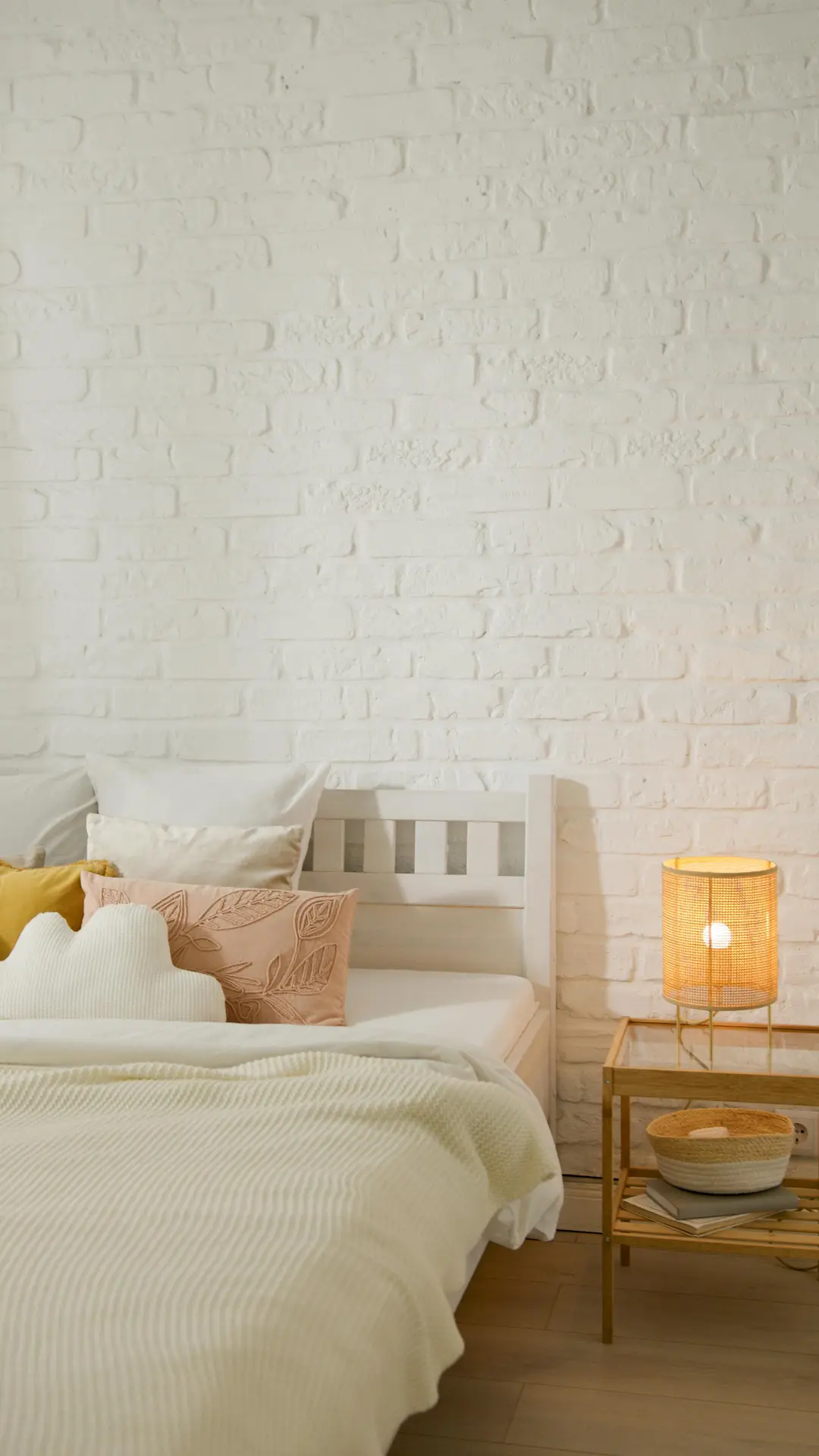 Spring decor ideas shown in a cozy bedroom with a white brick wall, layered neutral bedding, soft accent pillows, and a wooden nightstand topped with a warm-glowing rattan bedside lamp that adds natural texture and relaxed charm.