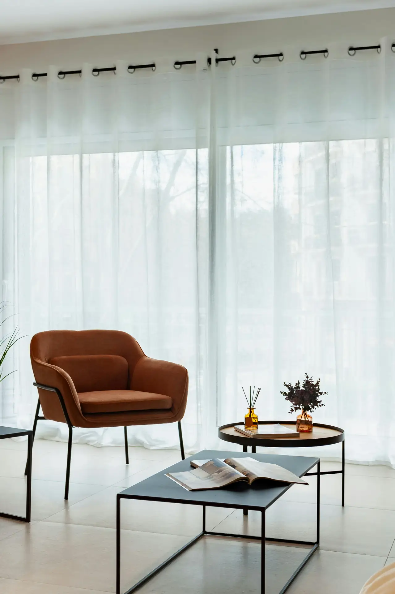 Bright living area with floor-to-ceiling sheer curtains filtering natural daylight, paired with a warm brown accent chair, slim black coffee tables, and simple decorative vases. The airy setup reflects spring decor ideas that focus on lightness, clean lines, and flexible styling for modern or minimalist interiors.