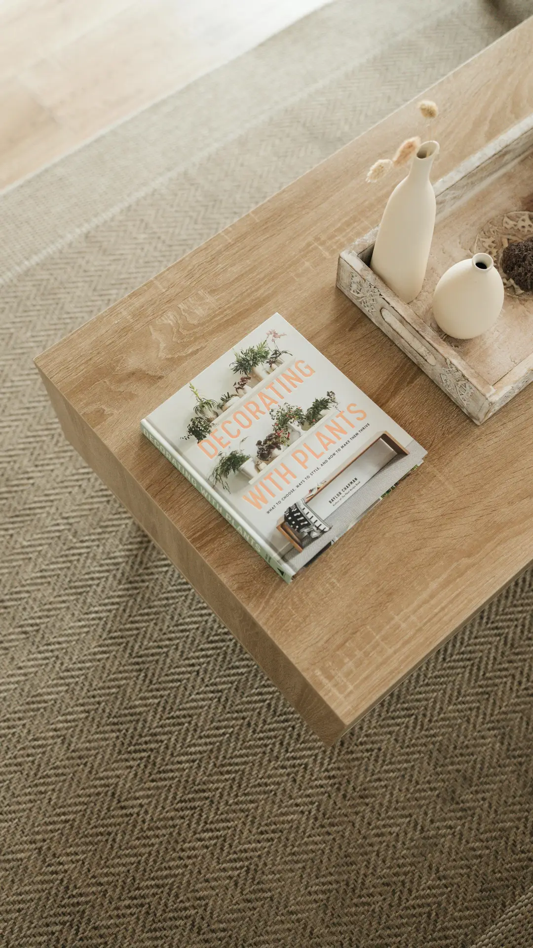 Spring decor ideas captured on a wooden coffee table featuring a spring themed coffee table book about decorating with plants, styled with neutral ceramic vases and a textured rug underneath, creating a calm and adaptable look for modern living spaces.