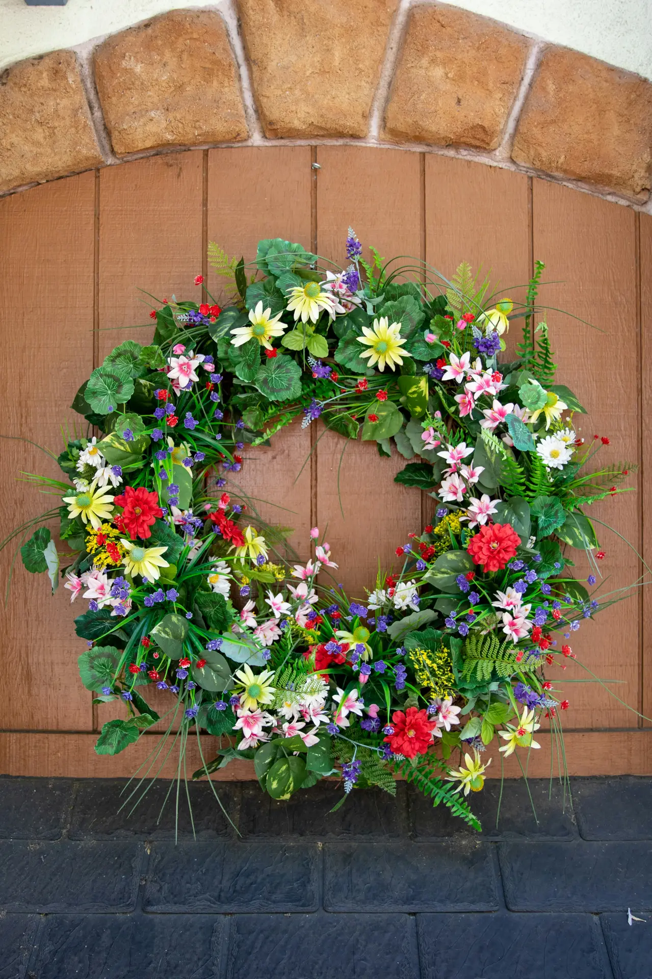 Spring decor ideas featuring a colorful spring wreath for front door styling, with a lush circular arrangement of green foliage and mixed flowers in soft pastels and bright tones, displayed against a wooden door framed by stone for a welcoming seasonal look.