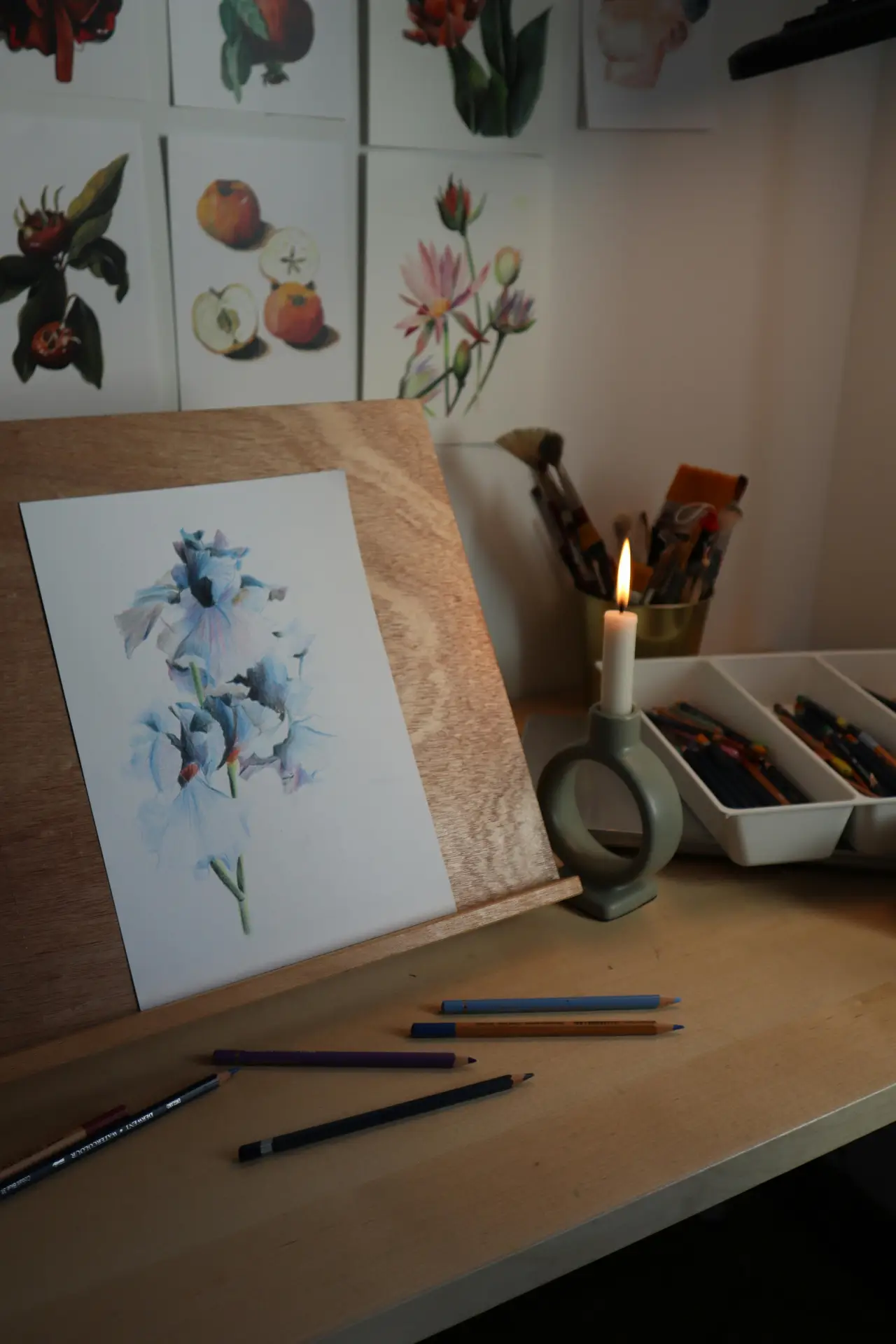Spring decor ideas shown through a cozy creative corner with watercolor prints, featuring a floral painting on a wooden stand, soft candlelight, art supplies on a desk, and botanical illustrations on the wall that add a calm, artistic touch to the space.