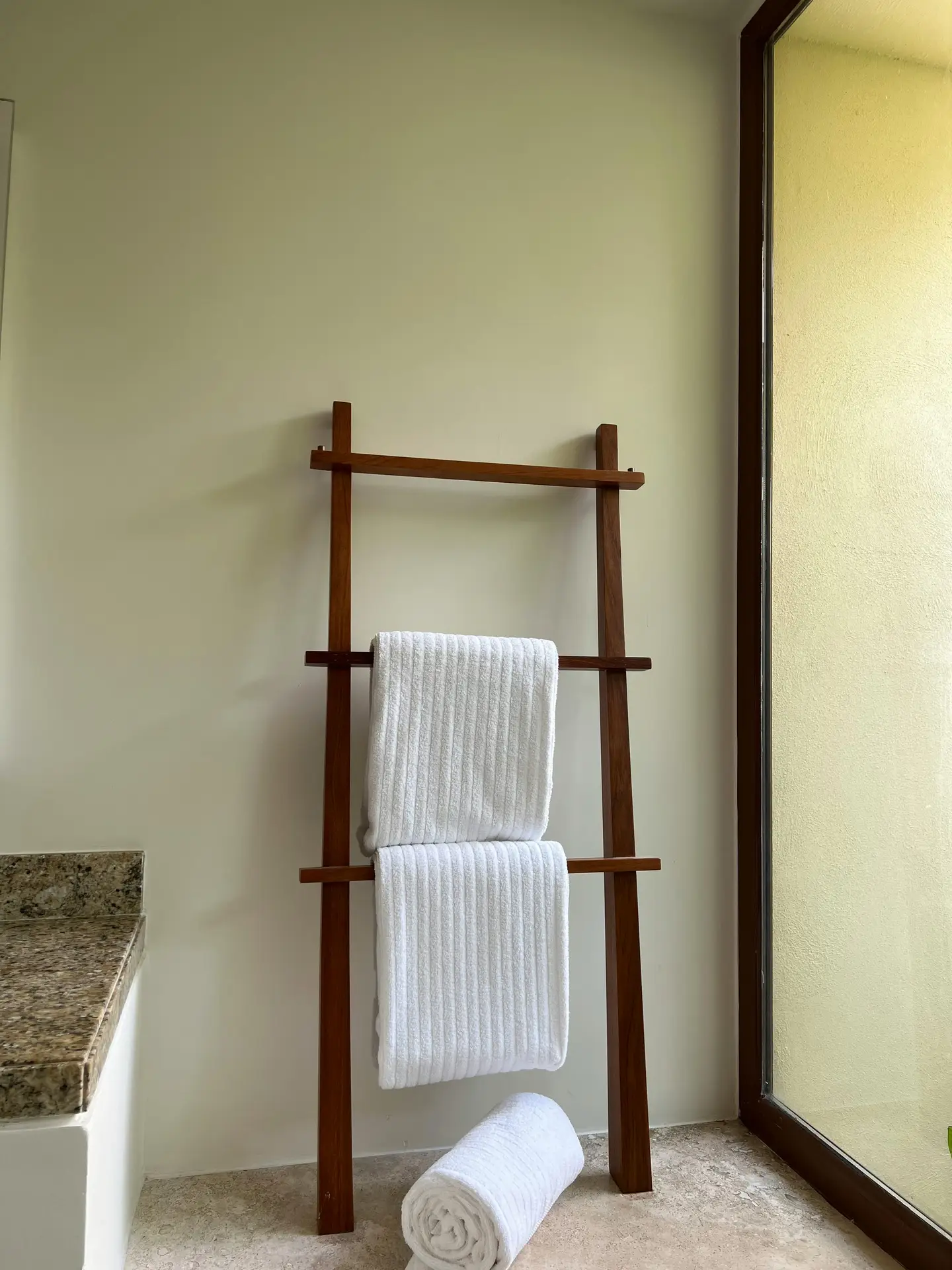 Spring decor ideas featuring white towels neatly draped over a wooden ladder rack in a bright bathroom, with soft natural light, neutral walls, a stone countertop edge, and a rolled towel on the floor creating a calm, spa-like atmosphere.