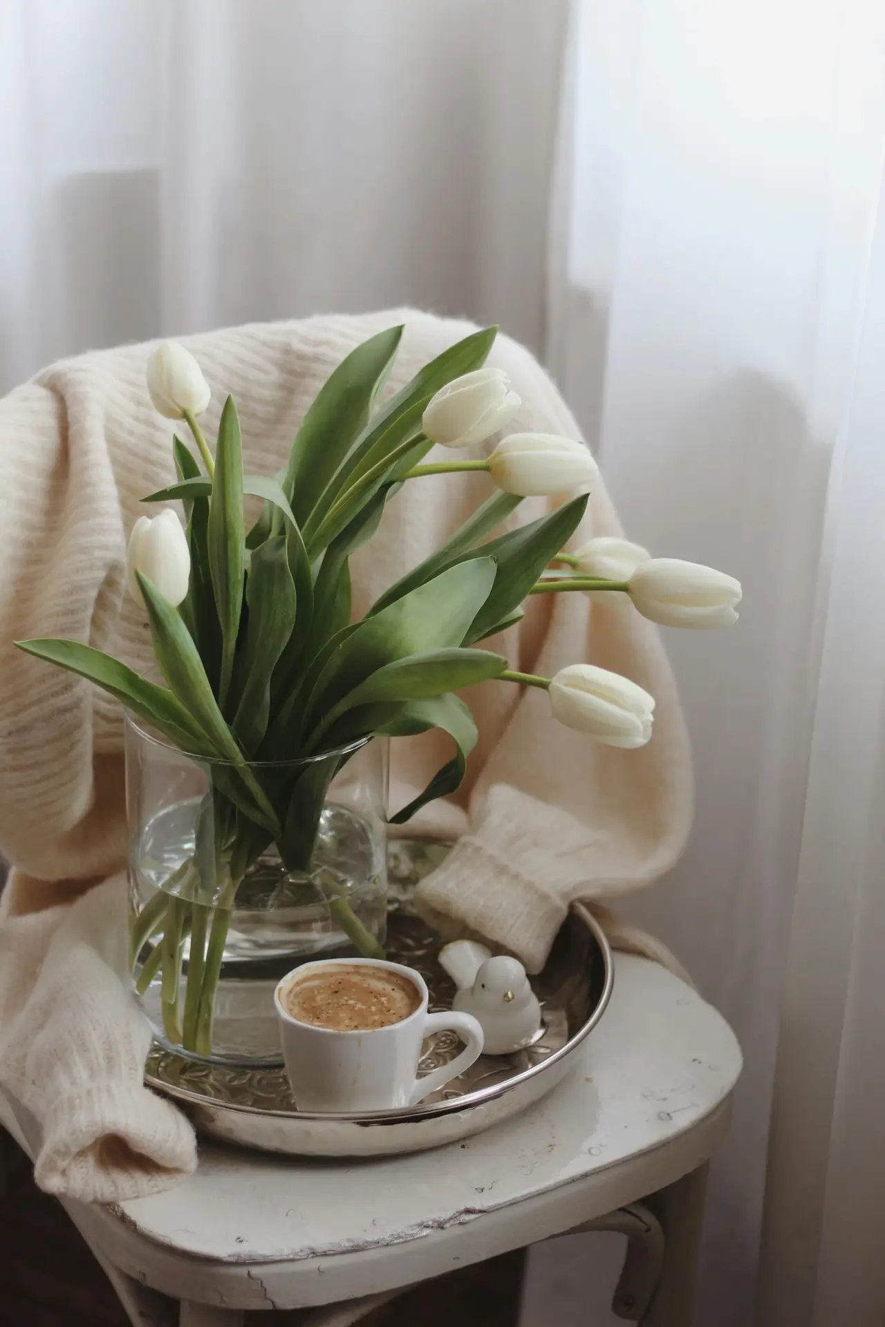A small round side table styled with a clear glass vase holding fresh white tulips, a soft knit throw draped over the chair, and a tray with a cup of coffee near a sheer curtain. This cozy setup reflects spring decor ideas that blend natural light, simple textures, and white tulips for a calm, versatile seasonal look.
