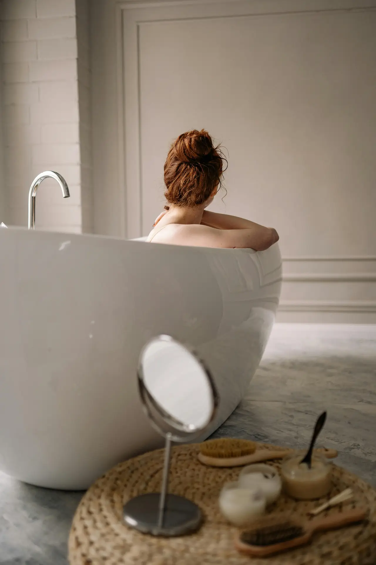 A calm bathroom scene showing a person relaxing in a smooth white acrylic tub, with a small tray of bath accessories in the foreground and soft neutral walls in the background, reflecting cozy bathroom ideas with a tub that focus on comfort, simplicity, and a spa-like atmosphere.